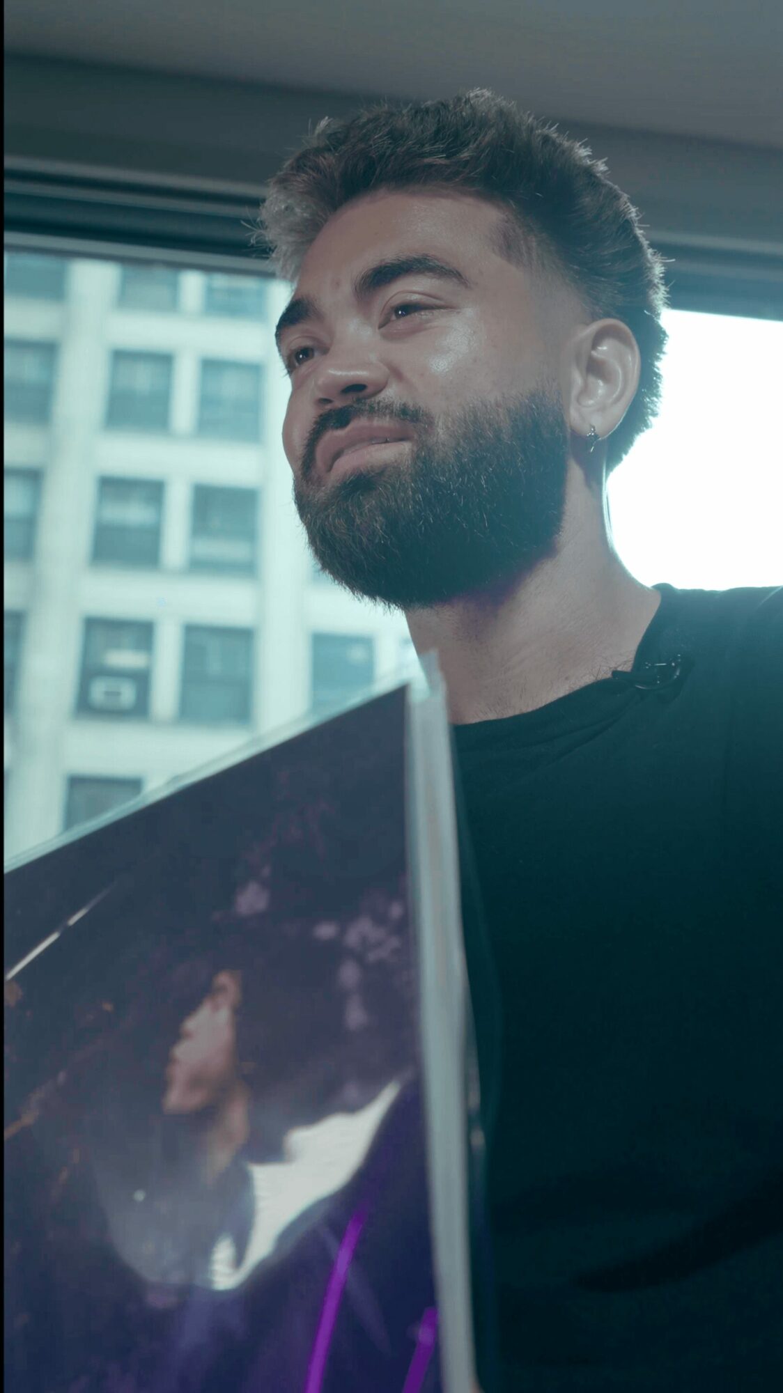 Young man with a beard looking out a window with a cityscape background, holding a tablet device.