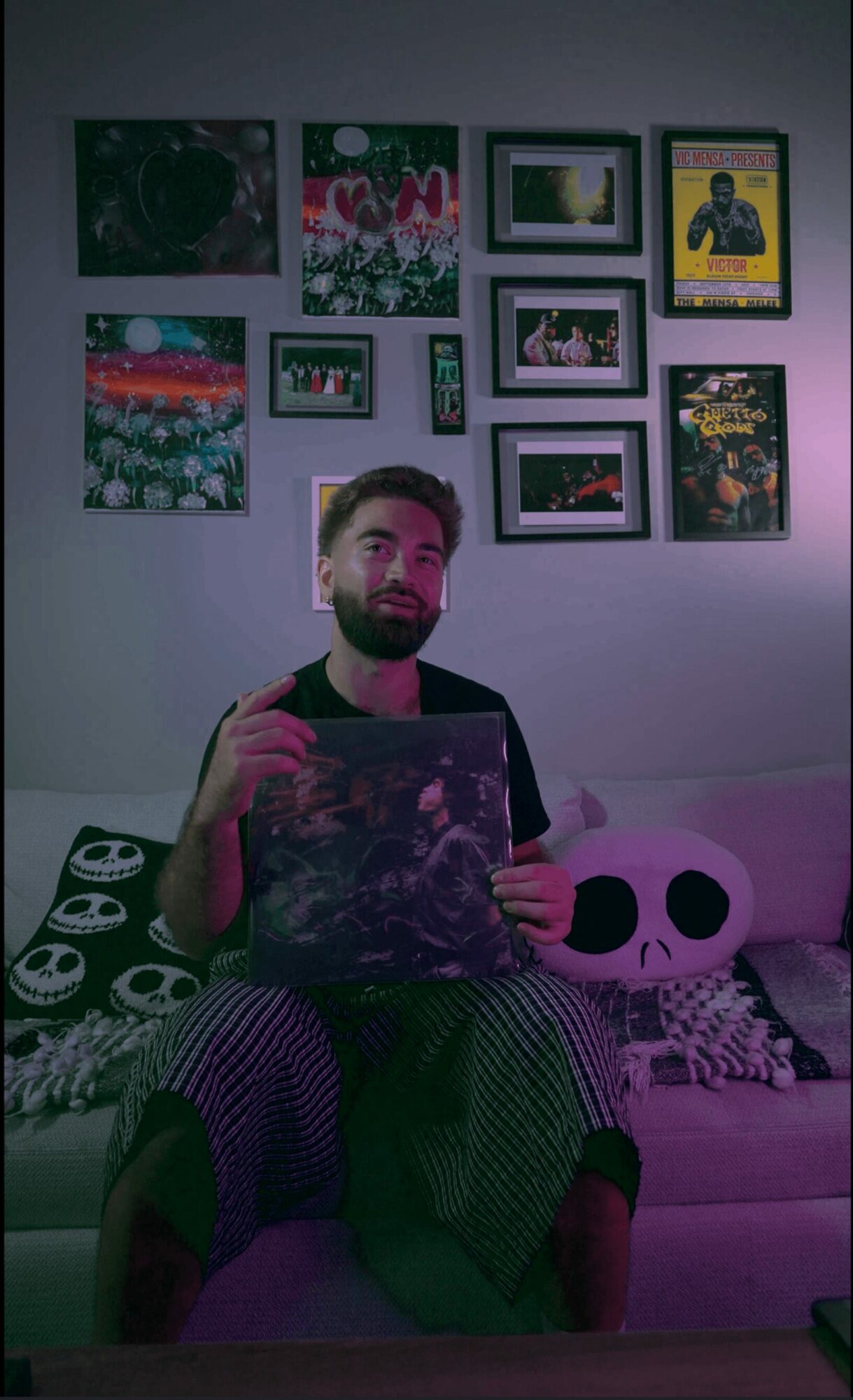 Person with beard sitting on a couch holding a vinyl record, surrounded by framed pictures and a panda pillow.