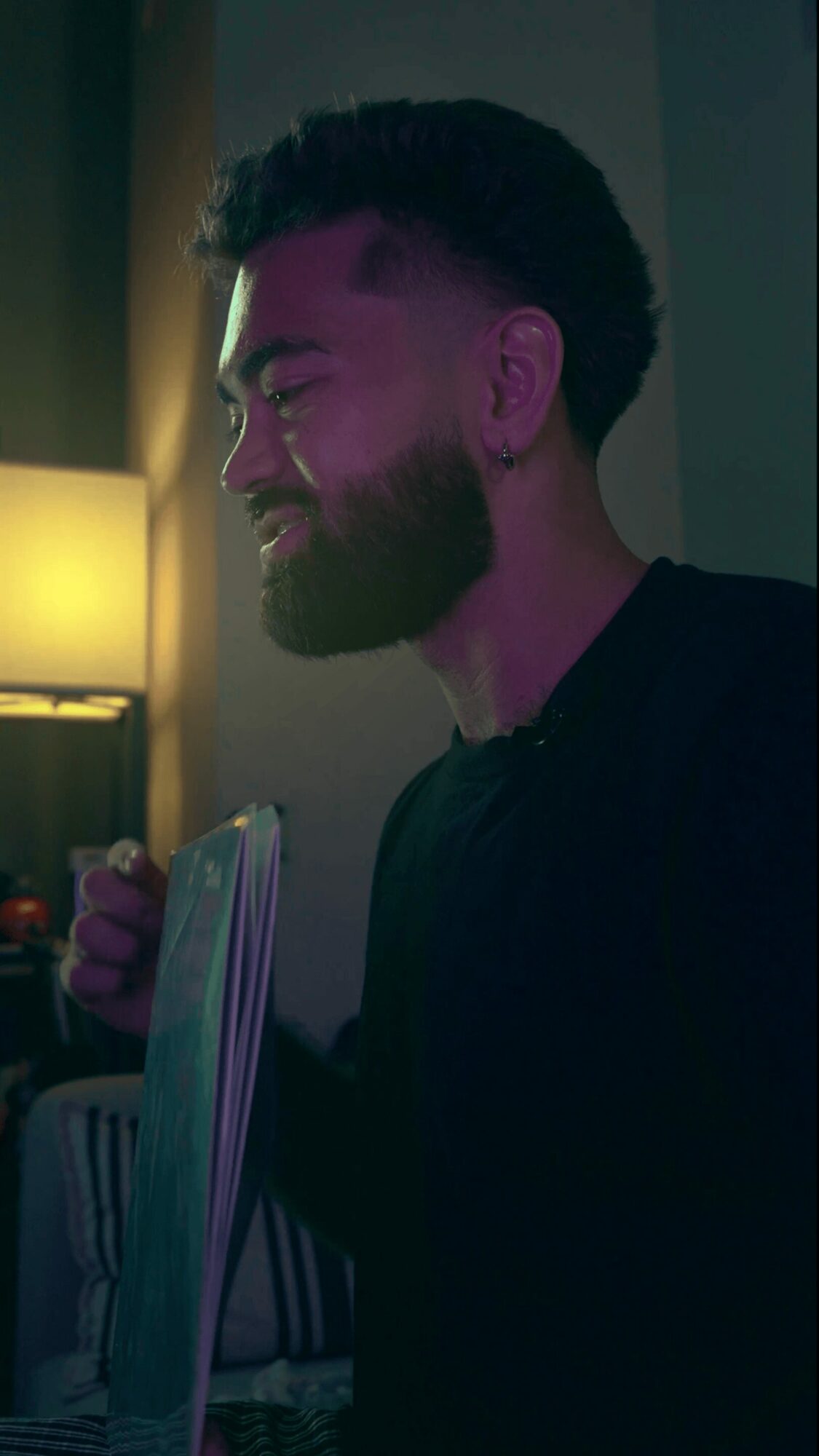 Person with a beard and short hair smiling, holding a book or magazine, in a dimly lit room with a lamp.