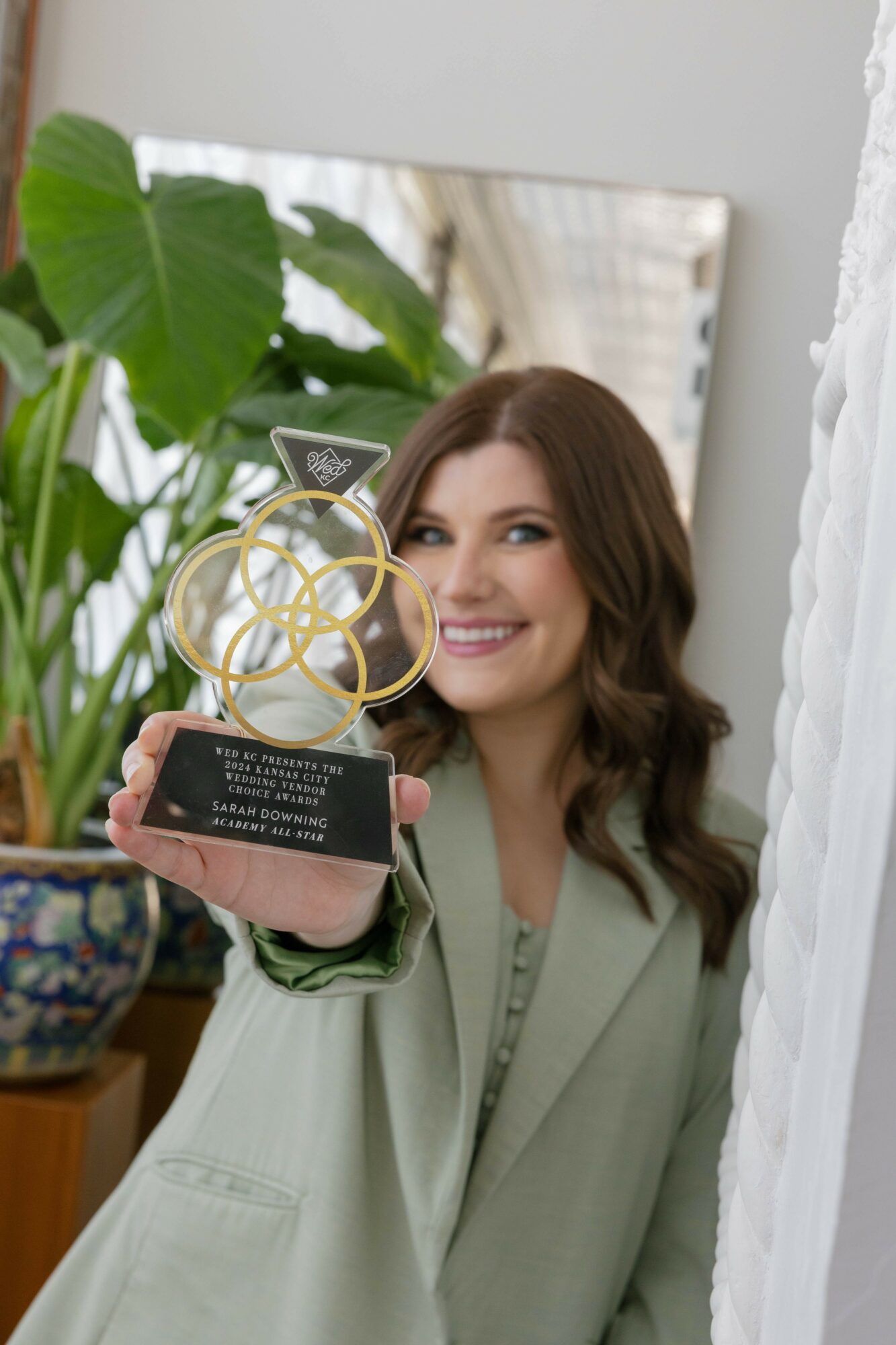 Woman with long brown hair smiling and holding a trophy in front of a mirror, with green plants in the background.