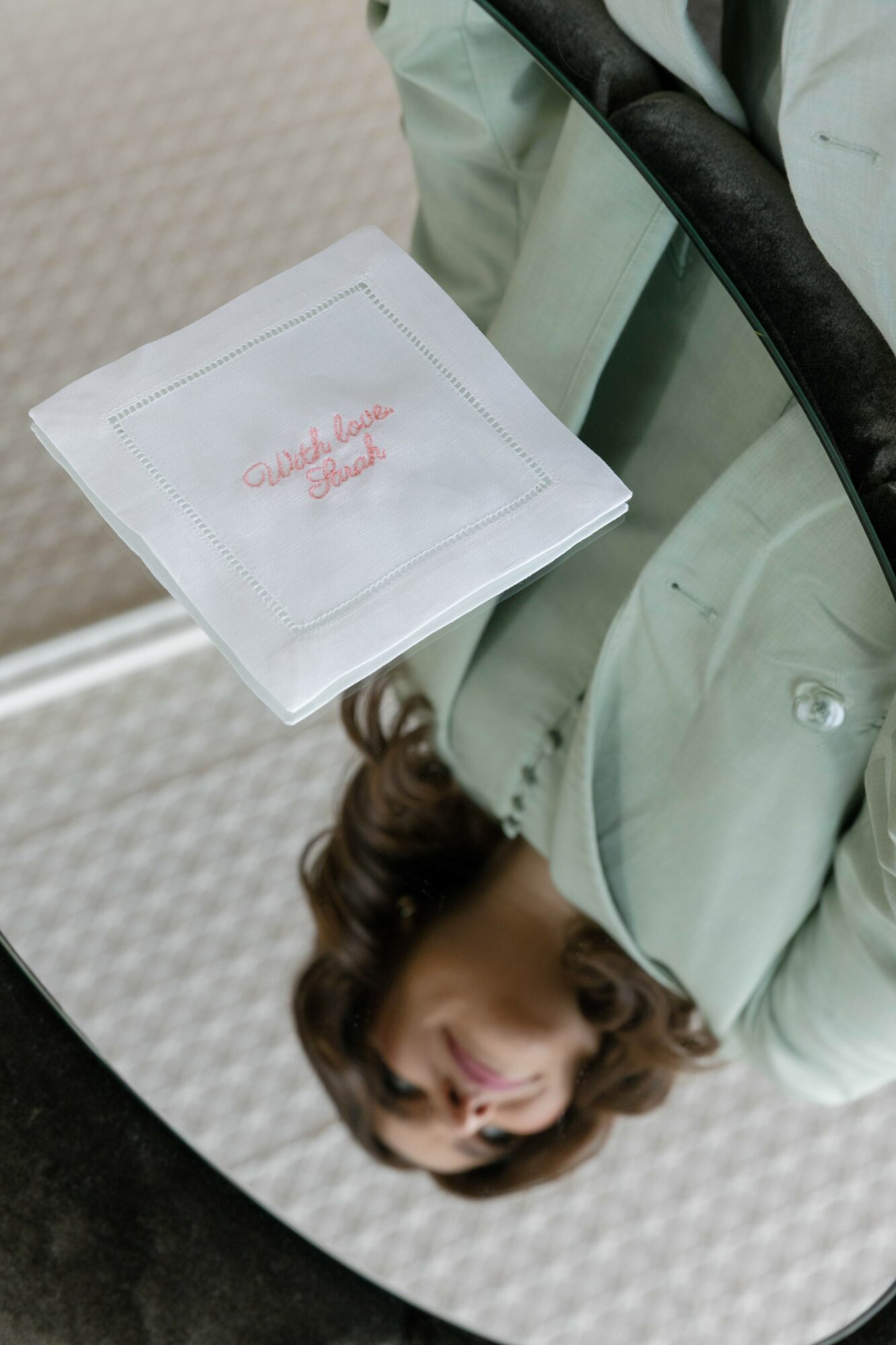 Person holding a white napkin with red embroidered text, reflected in a mirror, with a light-colored jacket and brown hair.
