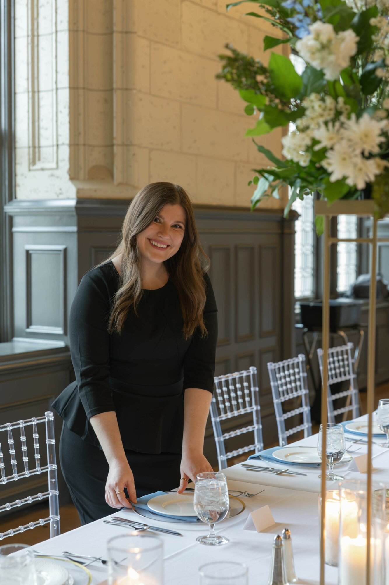 Woman with long hair in black dress standing at a decorated table in a bright room with large windows and floral arrangement.