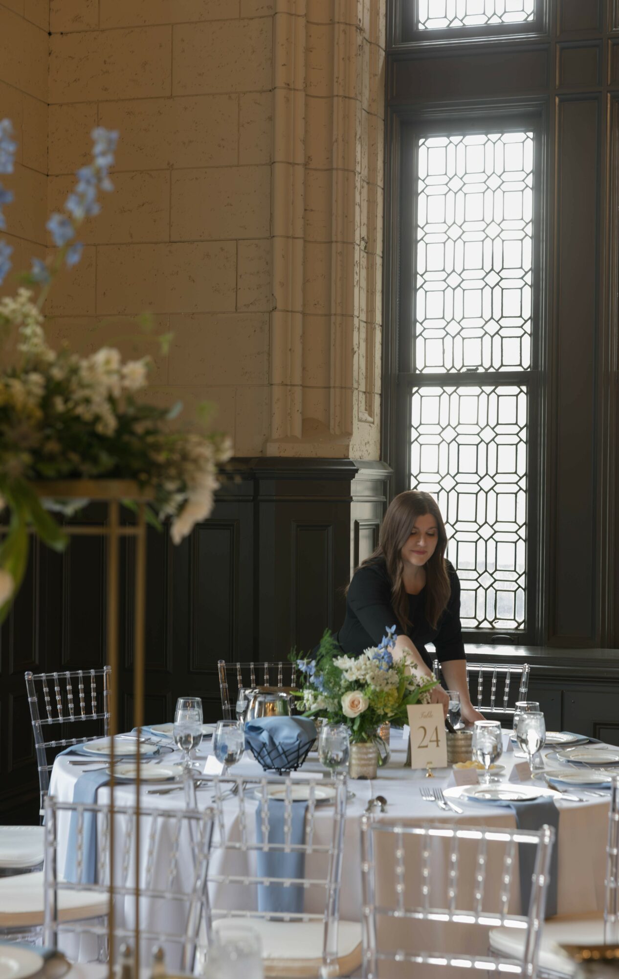 Woman arranging a table with flowers and glassware in a large room with tall windows and stone walls.