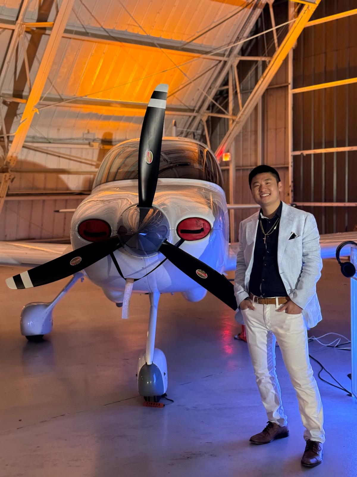 Young man in white jacket and jeans standing next to a small propeller airplane inside a hangar.
