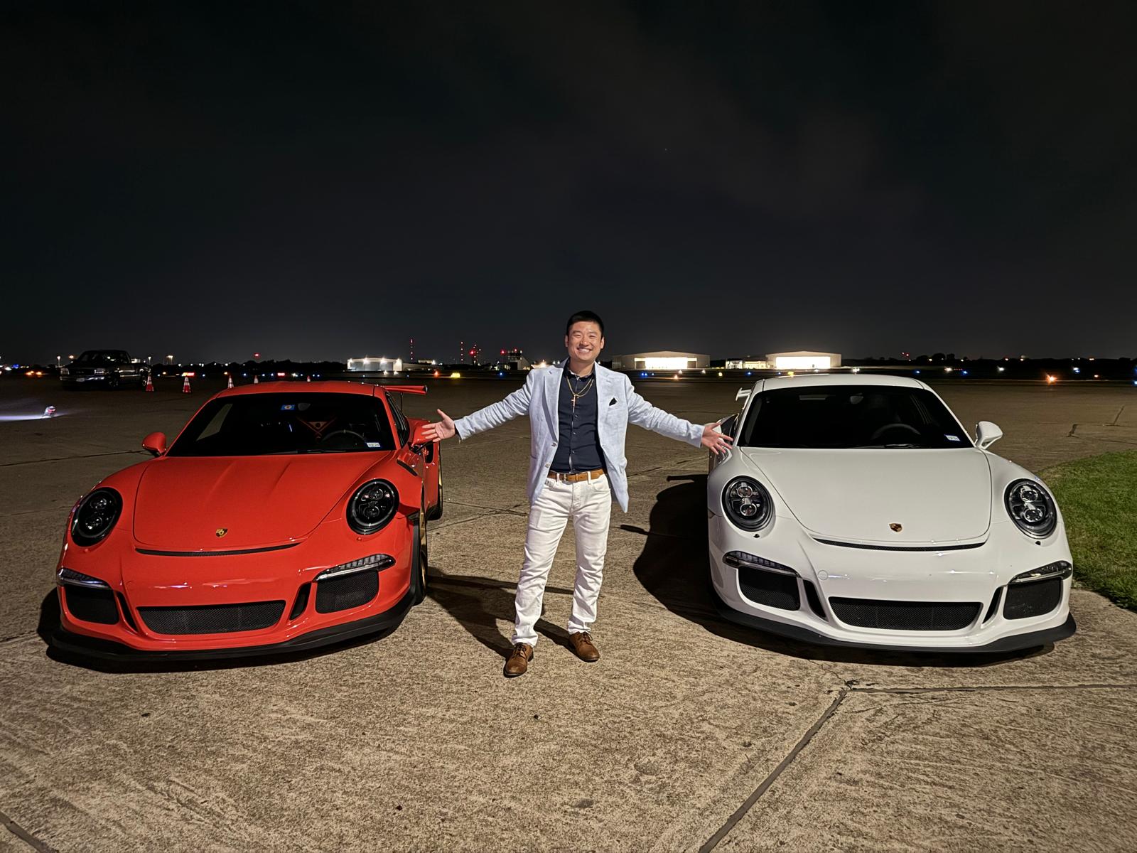 Person standing between two sports cars at night, one red and one white, with city lights in the background.