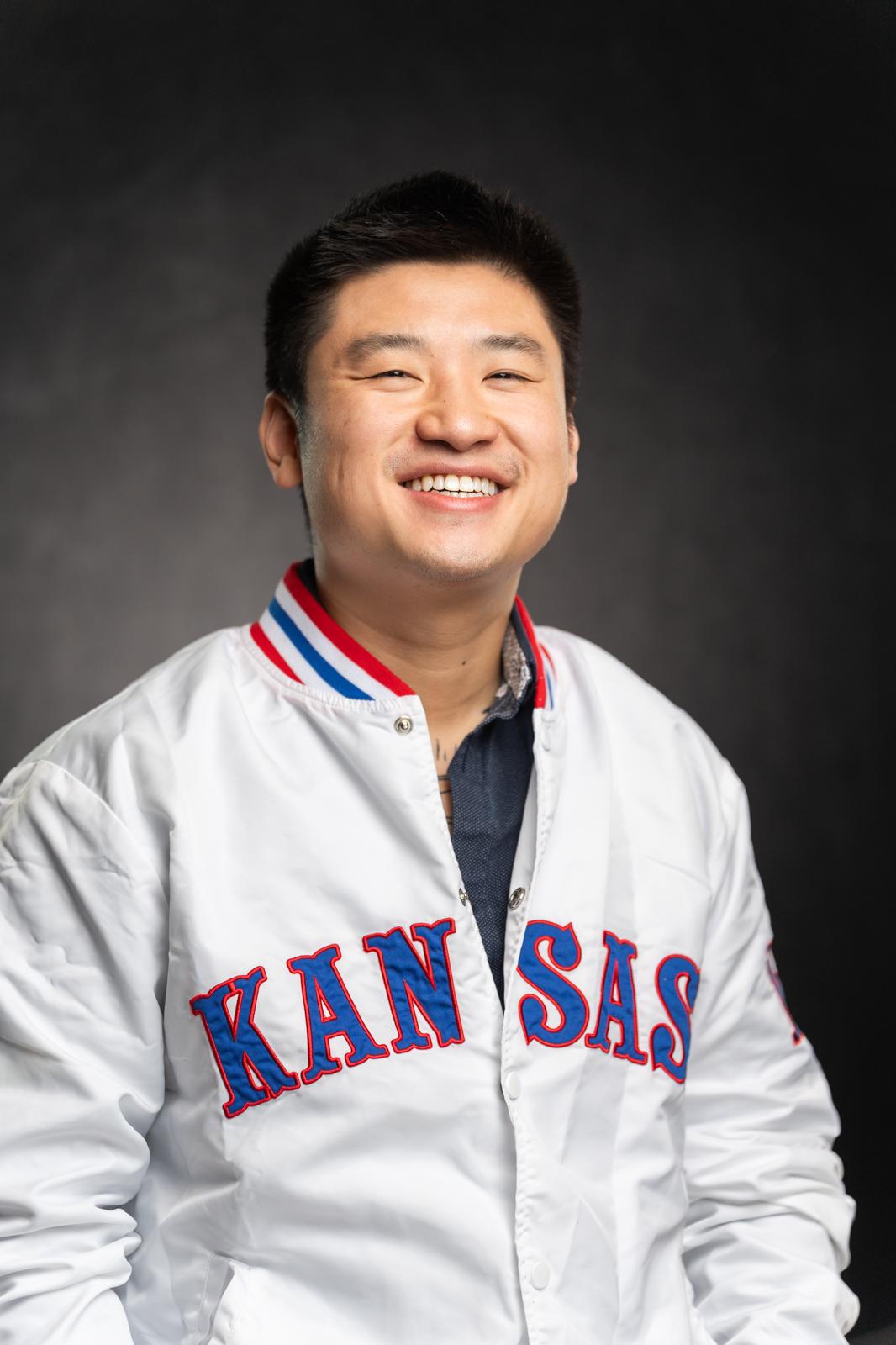 Smiling man wearing a white jacket with 'KANSAS' written on it, against a dark background.