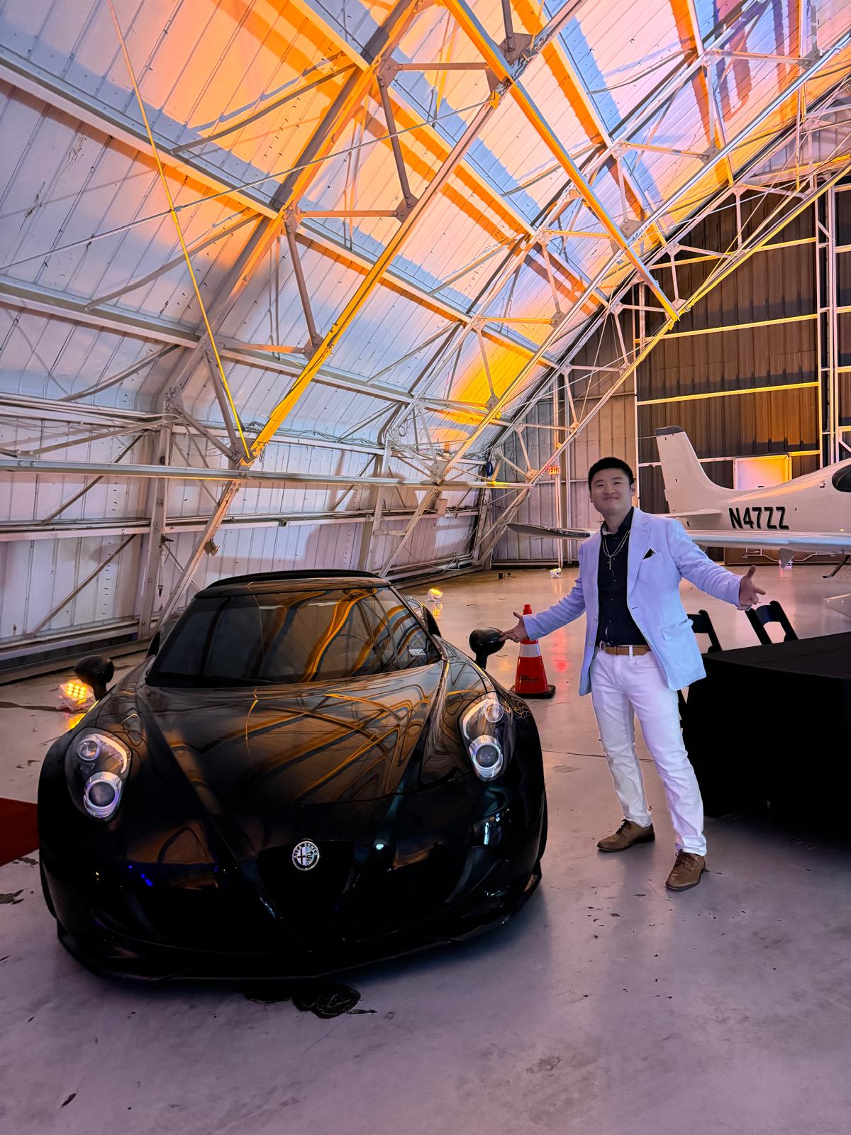 Person in white suit standing next to a black sports car inside a modern, glass-domed structure with a small aircraft in the background.