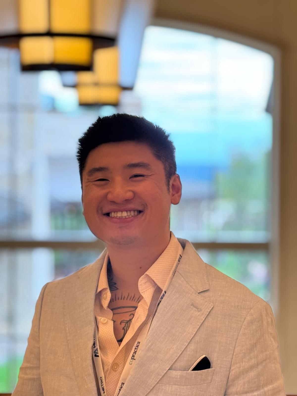 Smiling man in beige suit indoors with large windows and hanging lights in background.