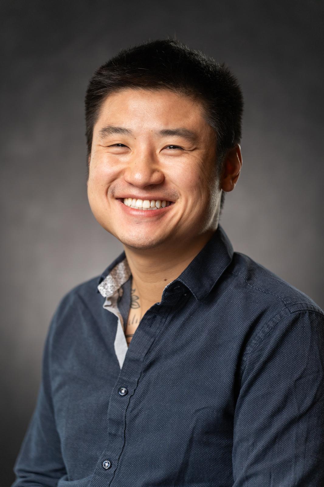 Smiling person with short black hair wearing a dark blue shirt against a dark background.