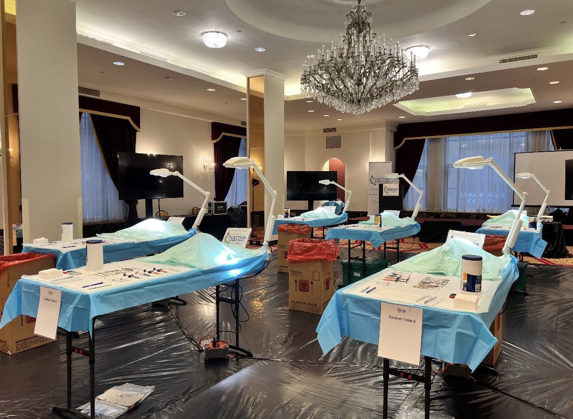Room set up for medical procedures with beds, lamps, and equipment, chandelier hanging from ceiling, dark curtains, and tables with supplies.