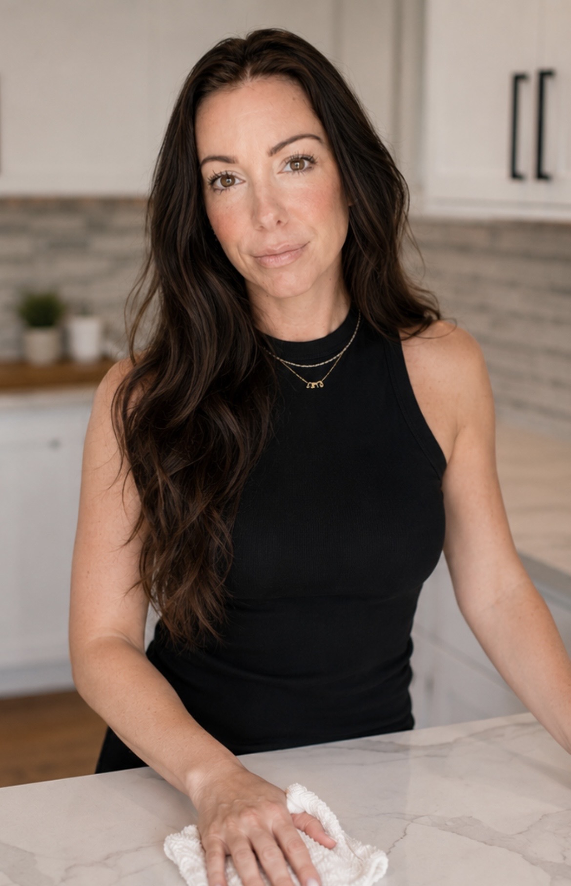 Woman with long dark hair wearing a black sleeveless top in a kitchen, looking at the camera.