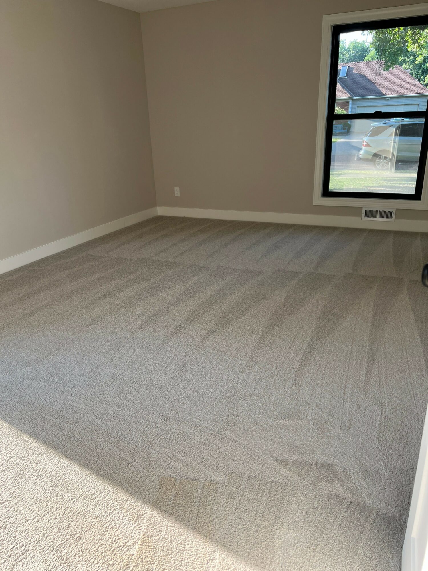 Empty room with window, beige walls, and light-colored carpeted floor, sunlight streaming in through window.
