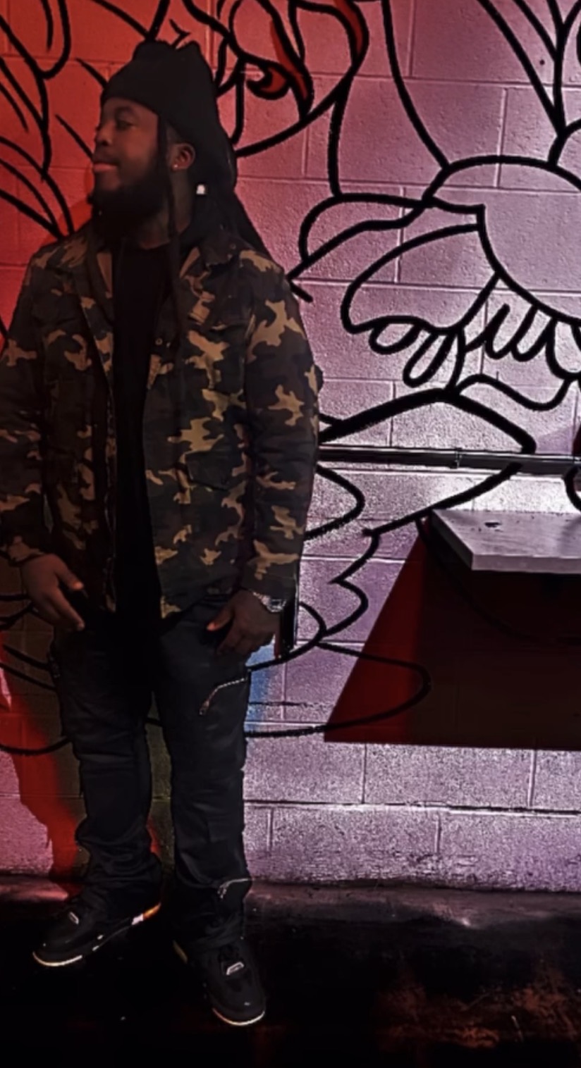 Man with a beard wearing a camouflage jacket and beanie, standing in front of a floral mural, with a table nearby.