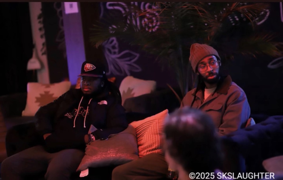 Two people sitting on a couch in a dimly lit room with colorful lighting, one wearing a beanie and glasses, the other in a hoodie and cap.
