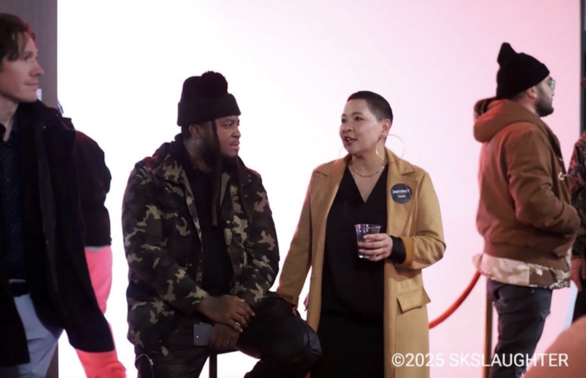 Four people standing in line, talking, with a pink and white background, one holding a drink, wearing casual clothing.