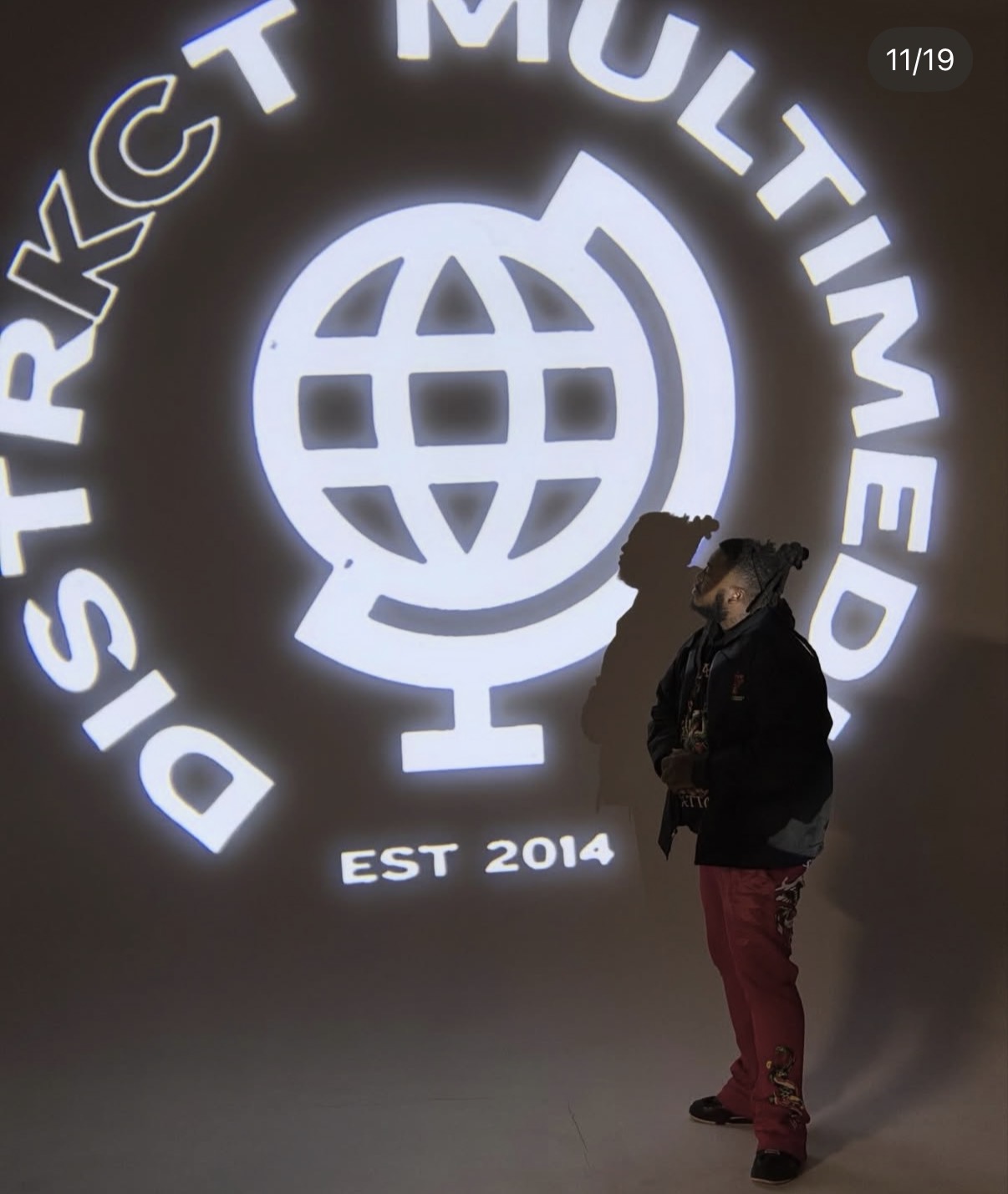 Person standing in front of a large illuminated logo with text and a globe symbol, with the person wearing a dark jacket and red pants.