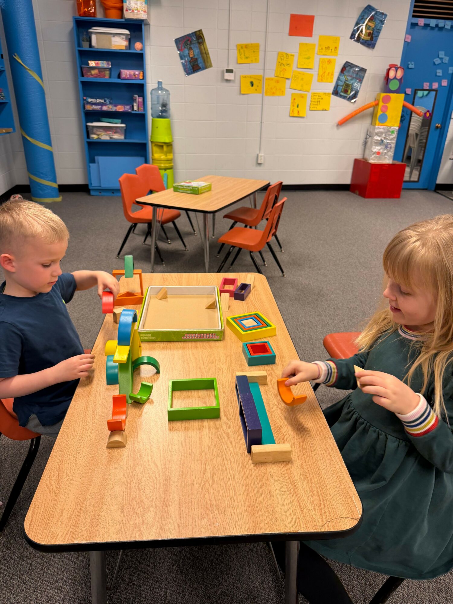 Child and adult playing with colorful wooden blocks on a table, with a whiteboard and storage shelves in the background.