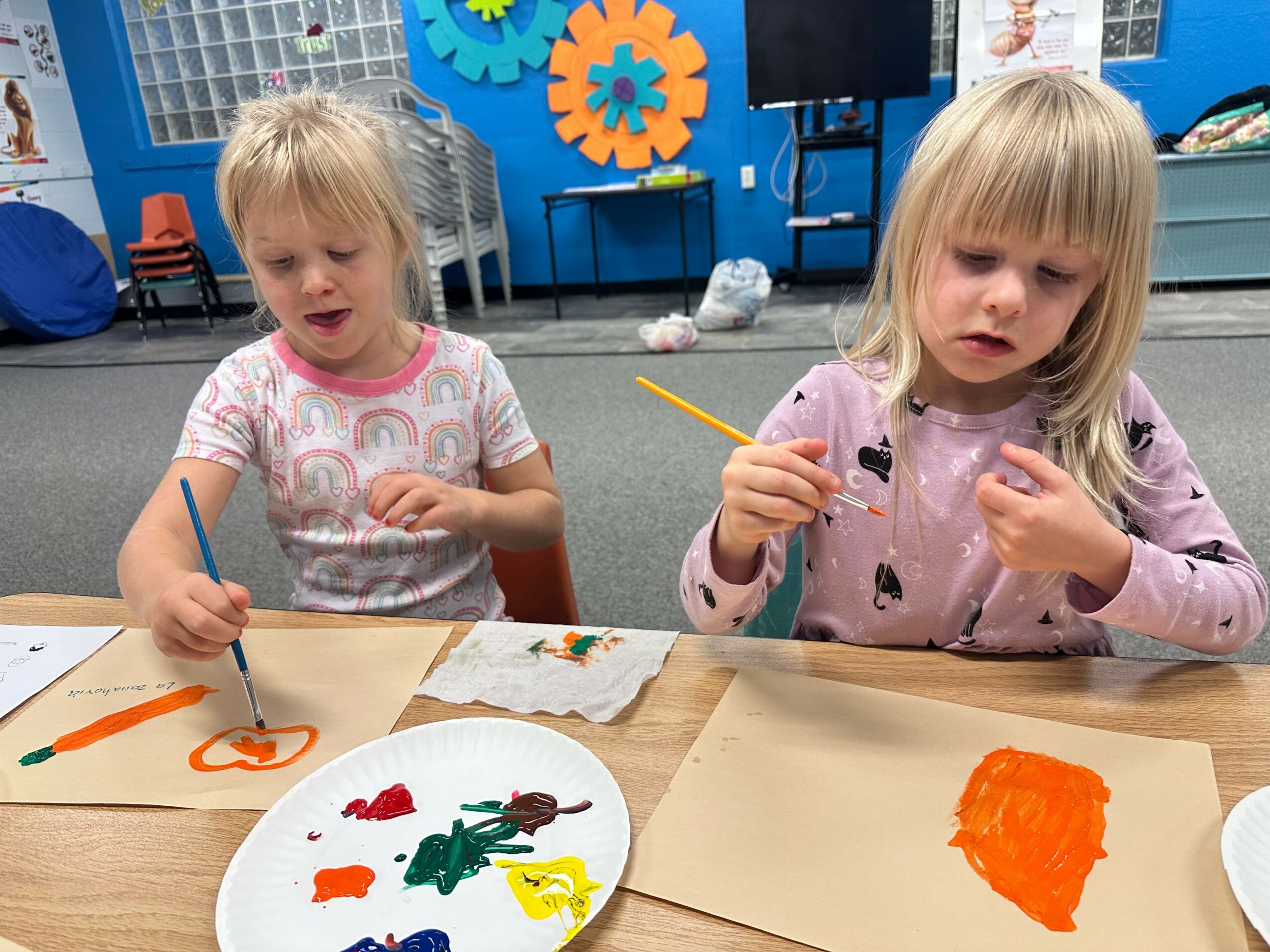 Two young girls sit at a table painting with brushes and colors, with art supplies and paper in front of them.