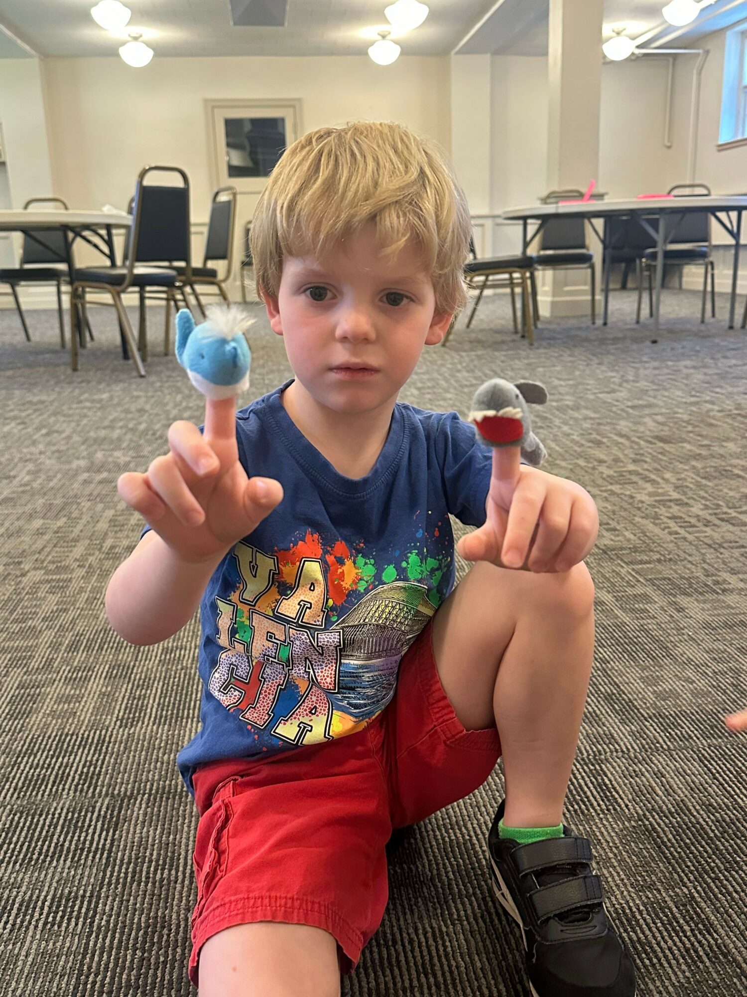 Young boy with blond hair sitting on a carpeted floor, holding a blue object, wearing a colorful shirt and red shorts.