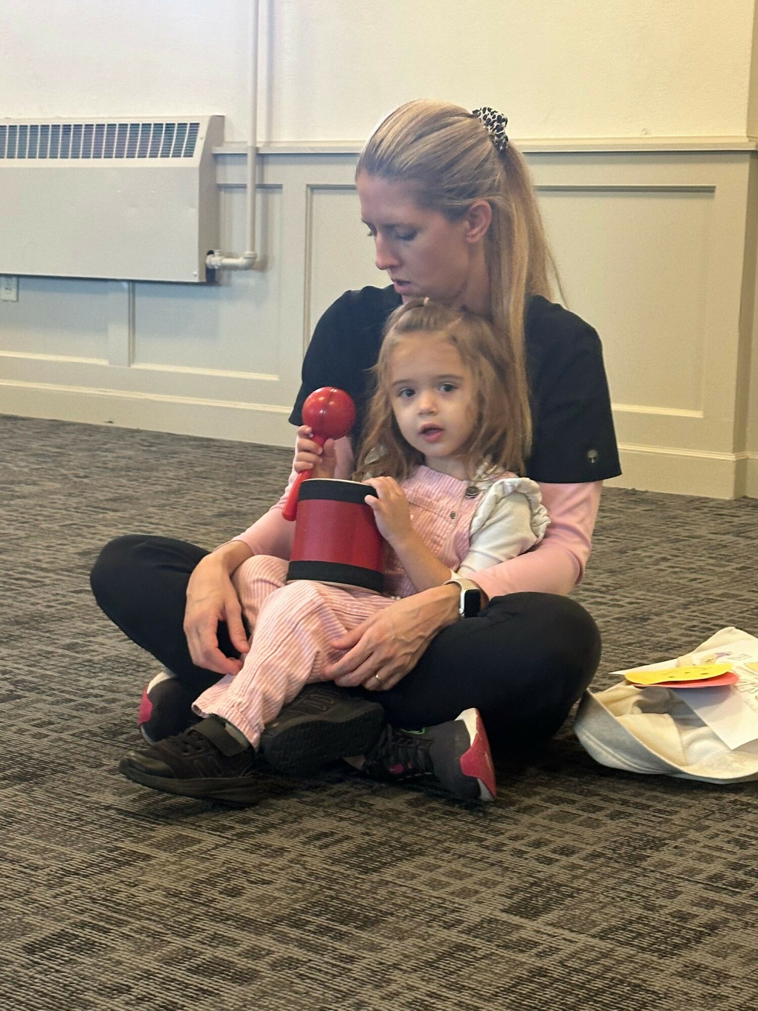 Adult woman sitting on the floor with a young girl, holding a red microphone, in a room with carpeted floor and white walls.