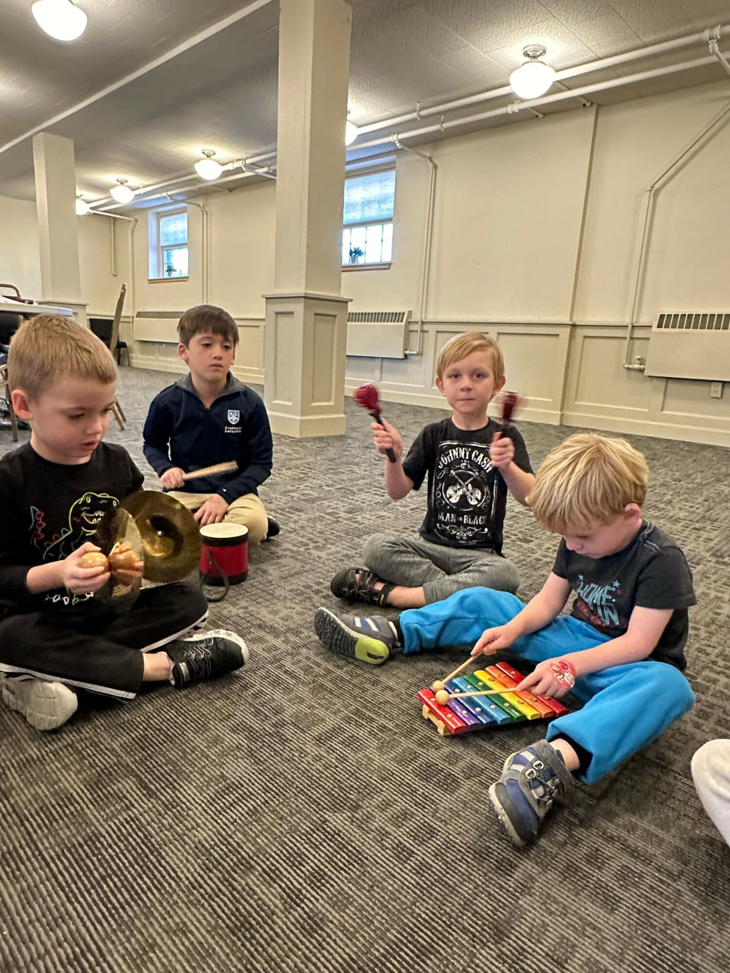 Three children sitting on the floor in a room, one standing, with books and a colorful bag, engaging in activities.
