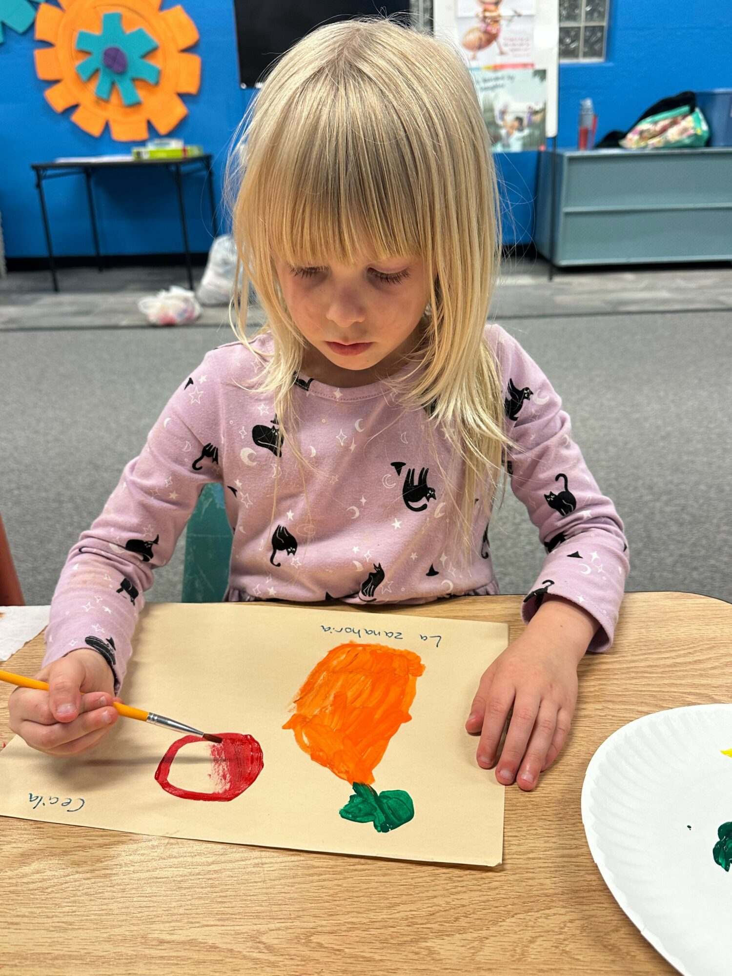 Child with blonde hair paints orange and green on a paper, sitting at a wooden table in a classroom.