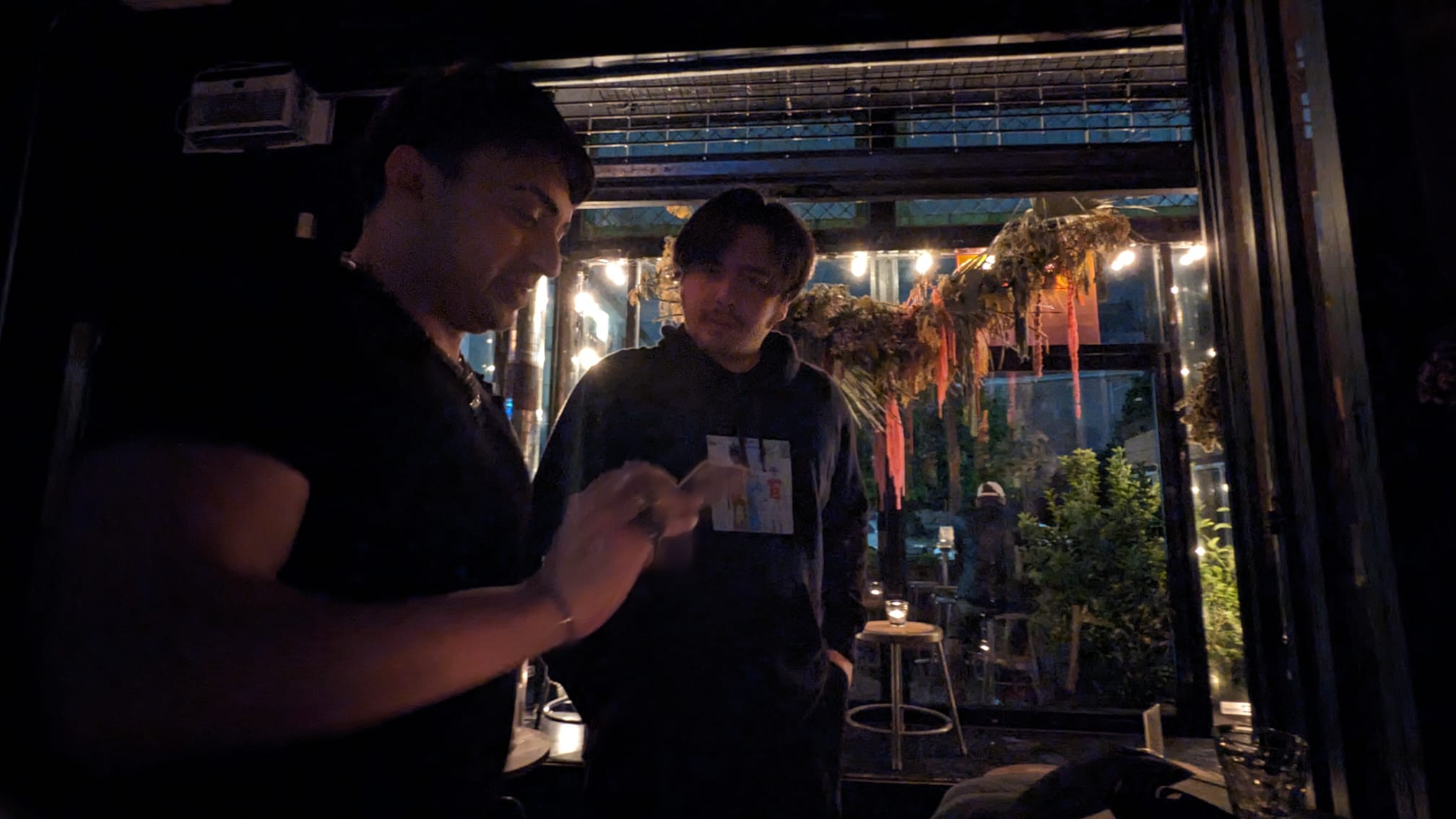 Two people standing indoors near a window with plants outside, one looking at a phone, the other holding a card.
