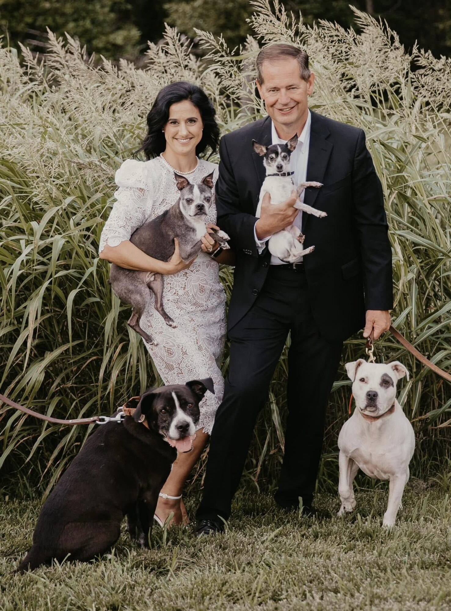 A man and woman stand outdoors with three dogs, surrounded by tall grass and plants, smiling at the camera.