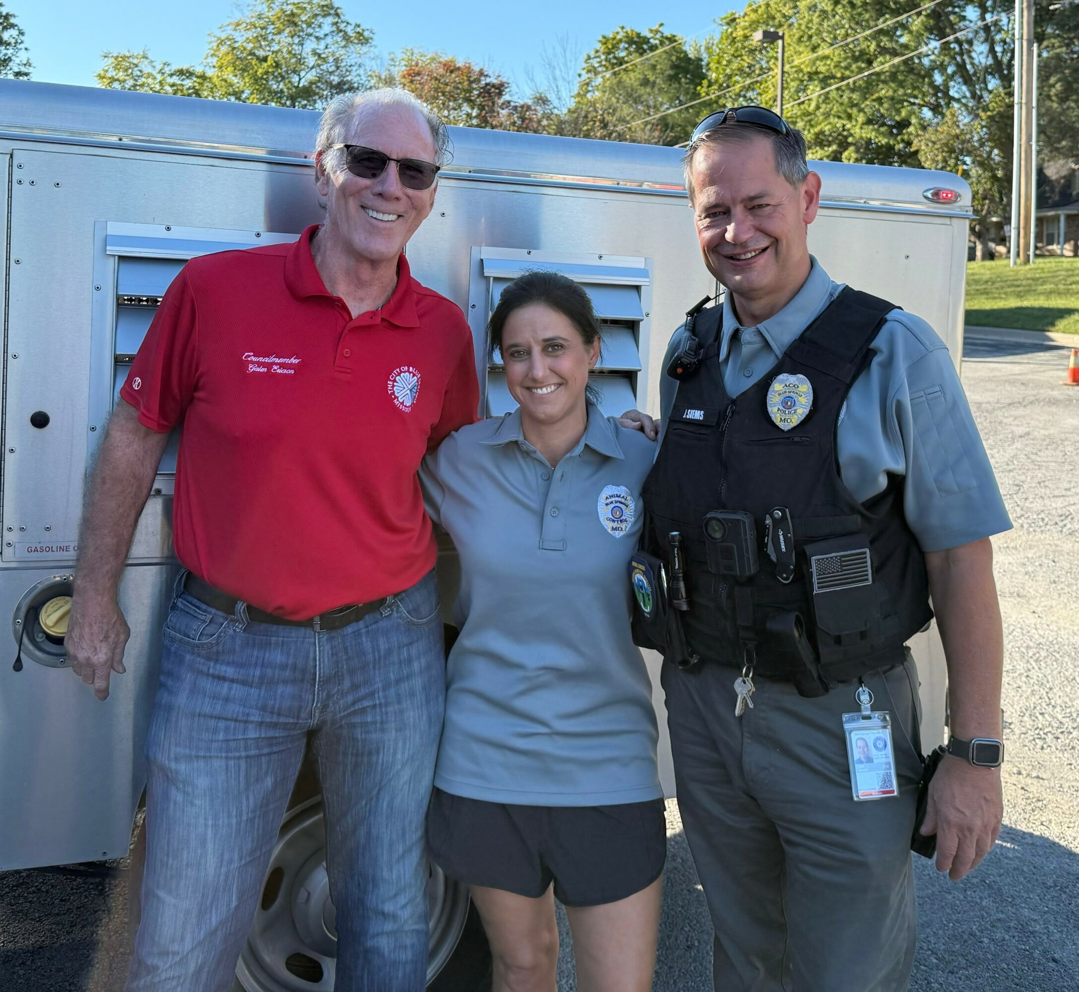 Three people standing outdoors in front of a vehicle, smiling, with trees and a blue sky in the background.