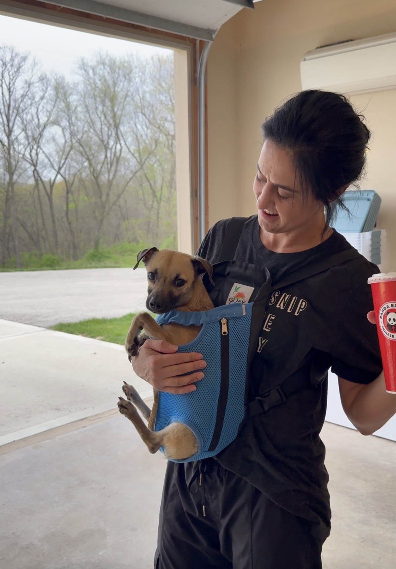Woman holding a small dog in a blue carrier, standing indoors near an open door with trees outside.