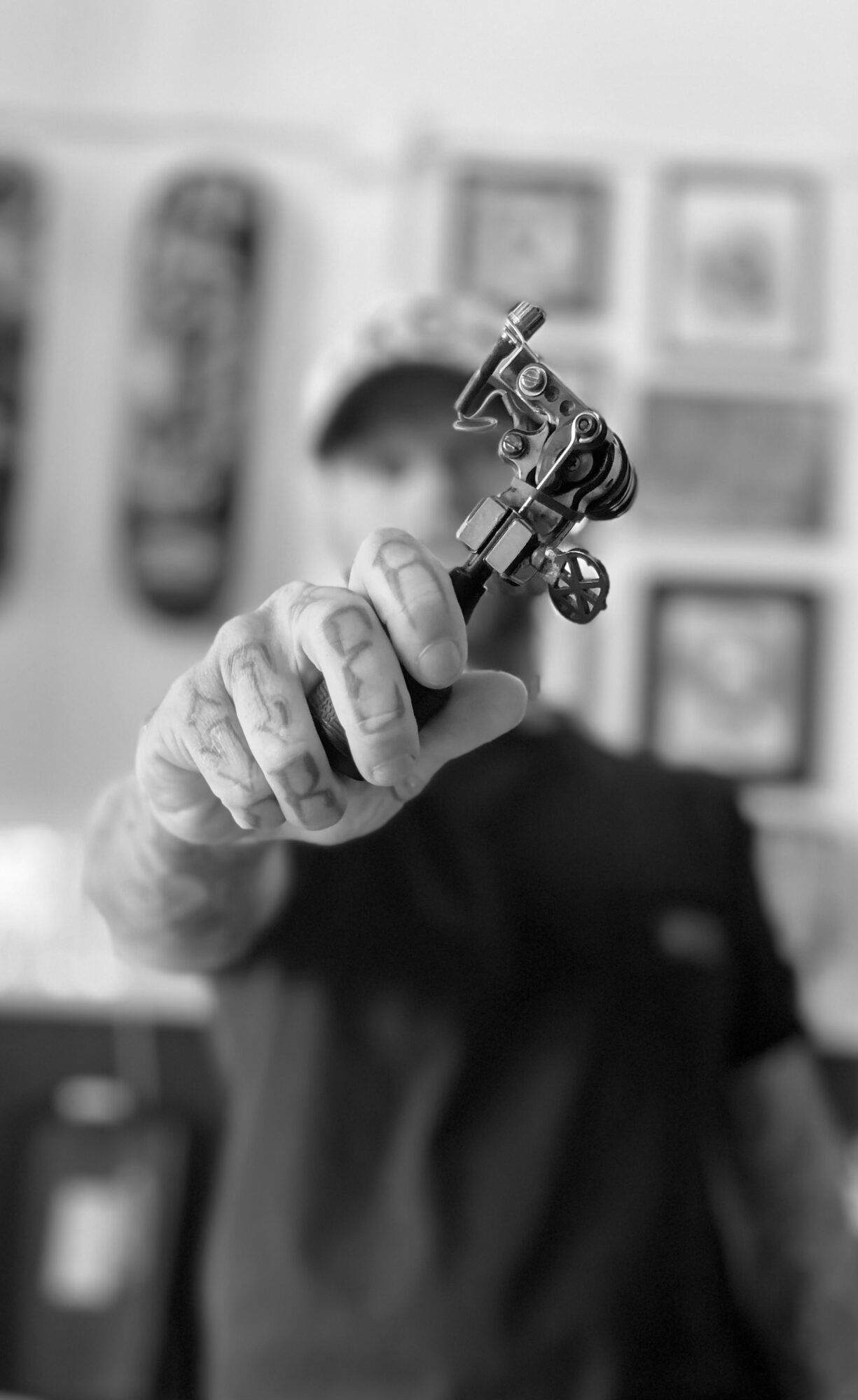 Person holding a tattoo machine, focus on hand and equipment, blurred background, black and white.