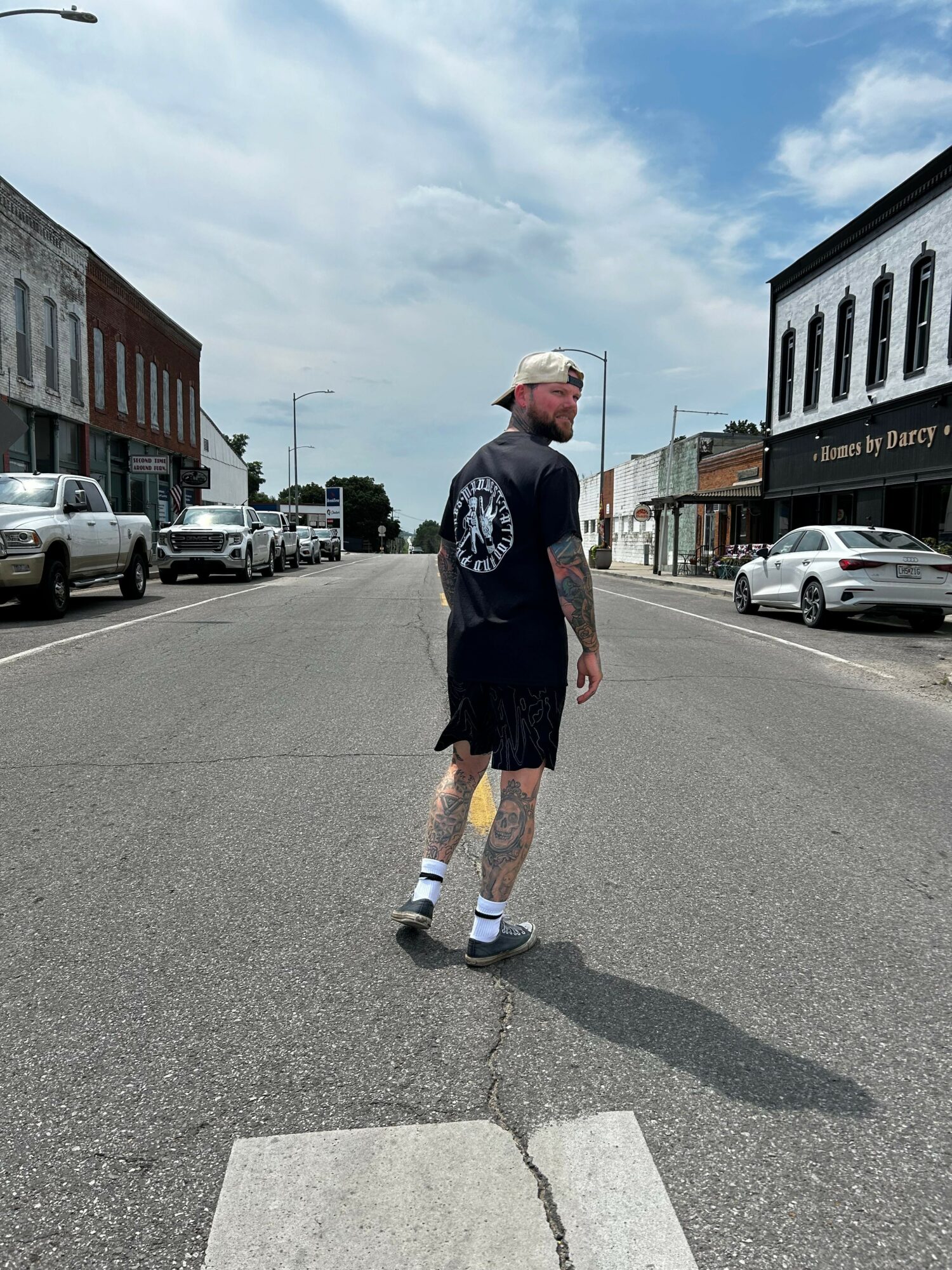 Person standing on street corner wearing a black shirt, shorts, and sneakers, with buildings and cars in the background.
