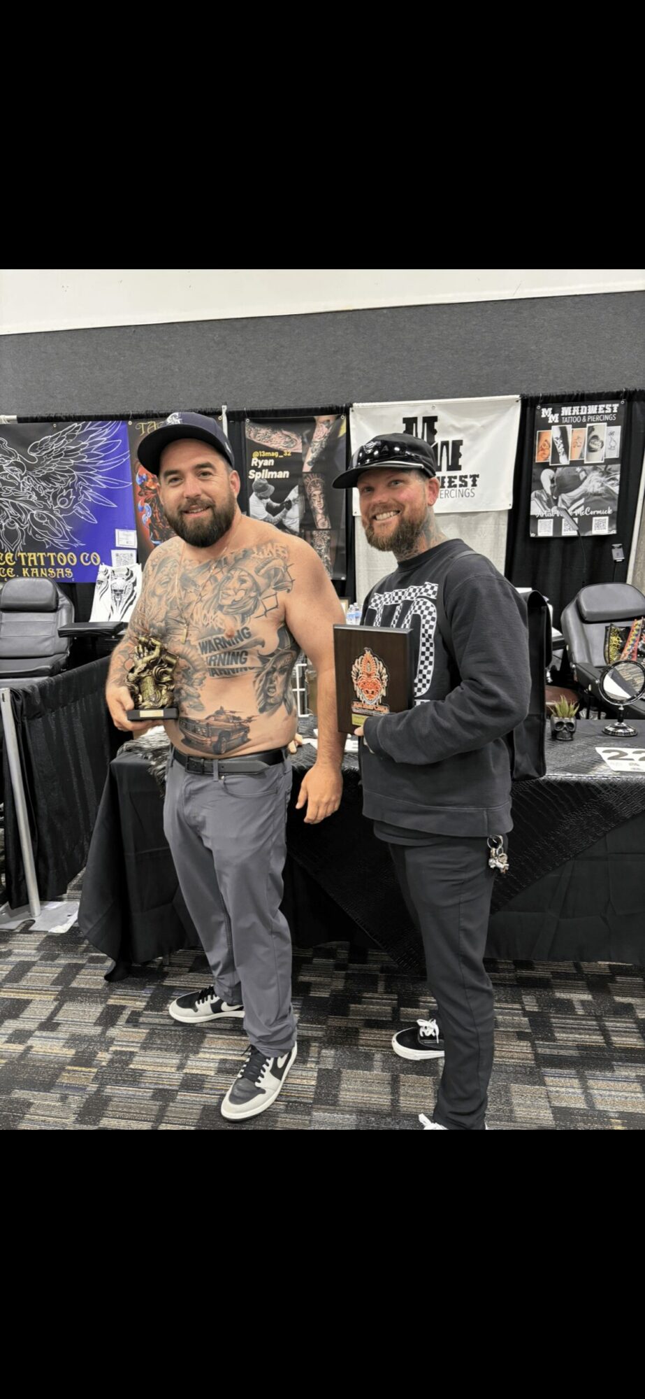 Two men standing indoors, one shirtless with tattoos and a cap, the other in a hoodie and glasses, smiling.
