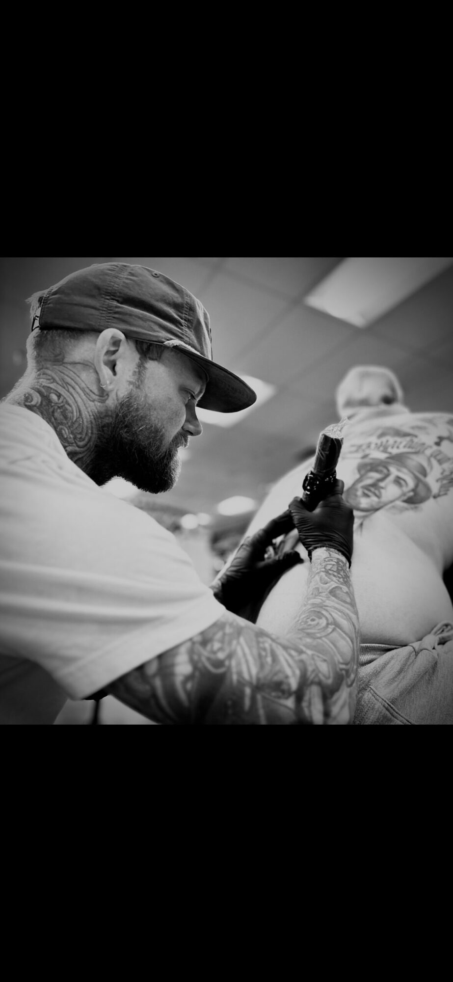 Tattooed man with a hat and beard, smiling, holding a tattoo machine, working on a person's arm in a tattoo studio.