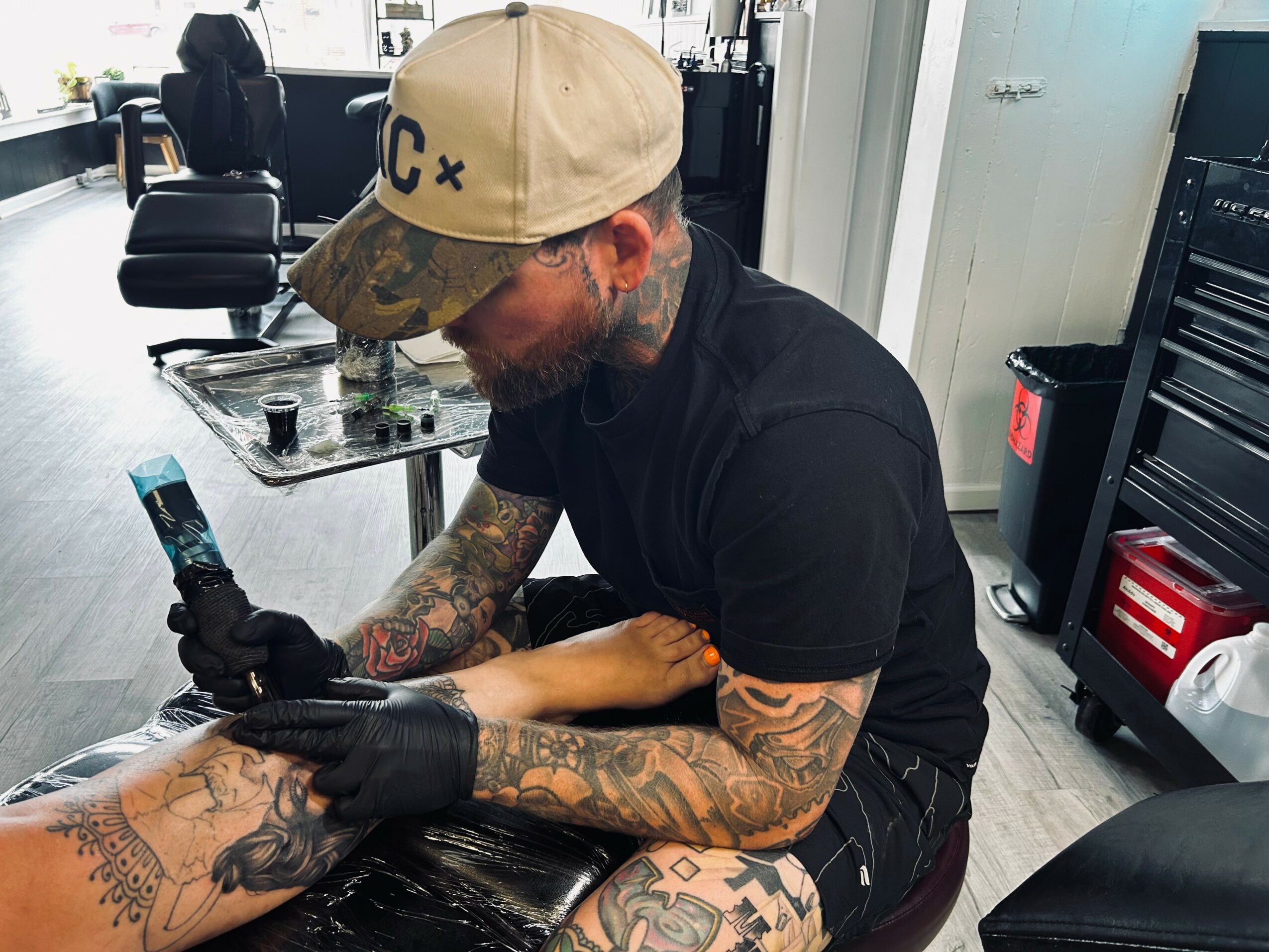 Tattooed man wearing a beige cap and black shirt getting a tattoo on his arm in a tattoo studio.