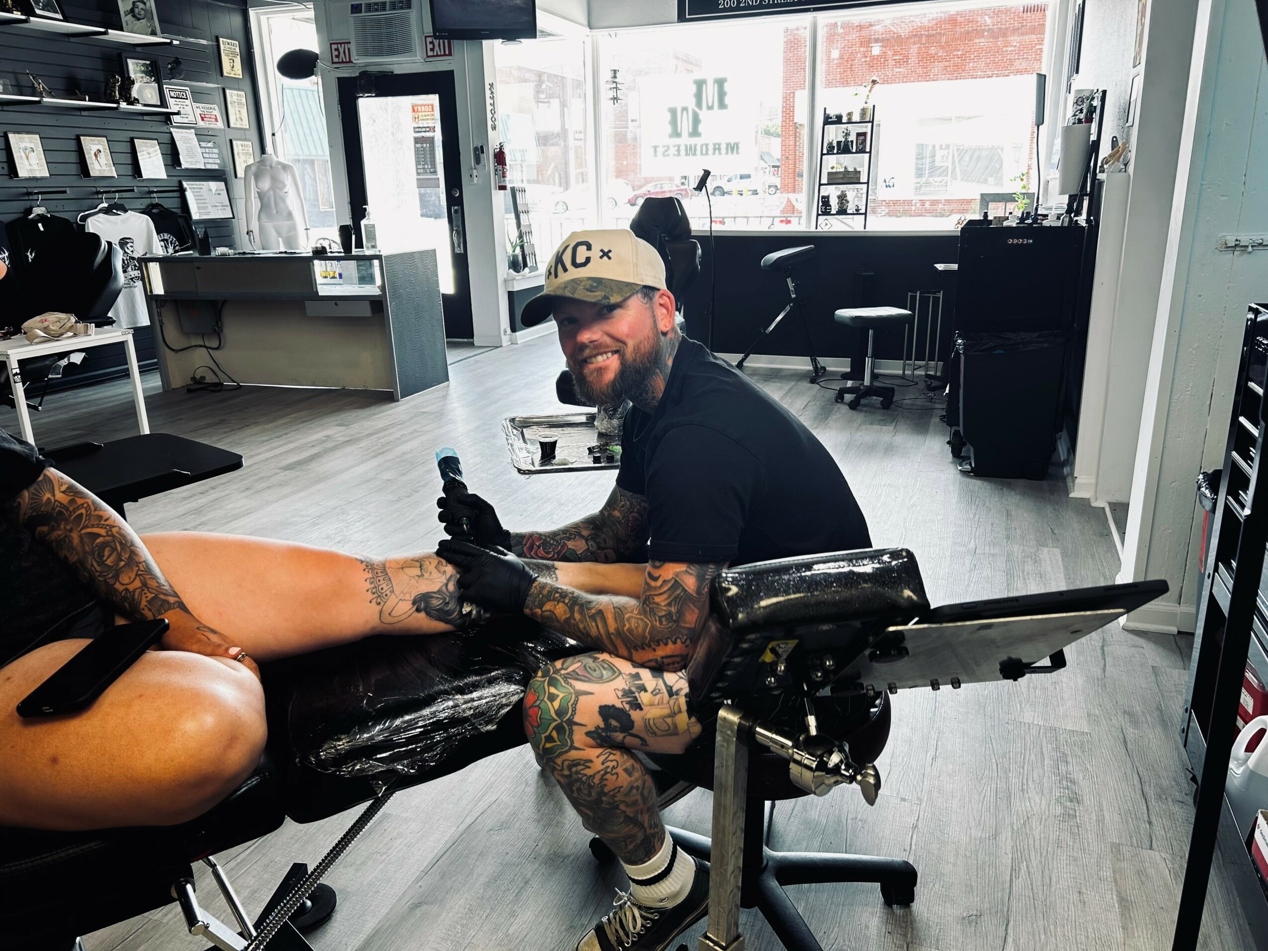 Tattoo artist working on a client's arm in a tattoo studio with large windows and equipment in the background.