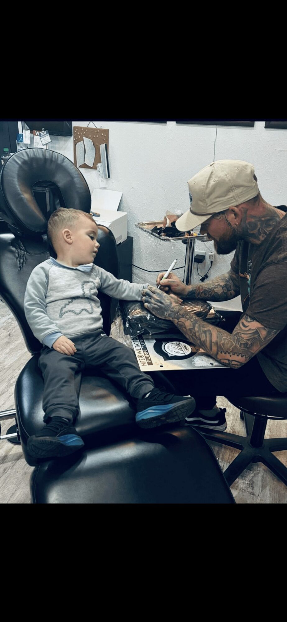 Child sitting in a chair, getting a tattoo from a tattoo artist in a studio, with equipment and chairs visible in the background.