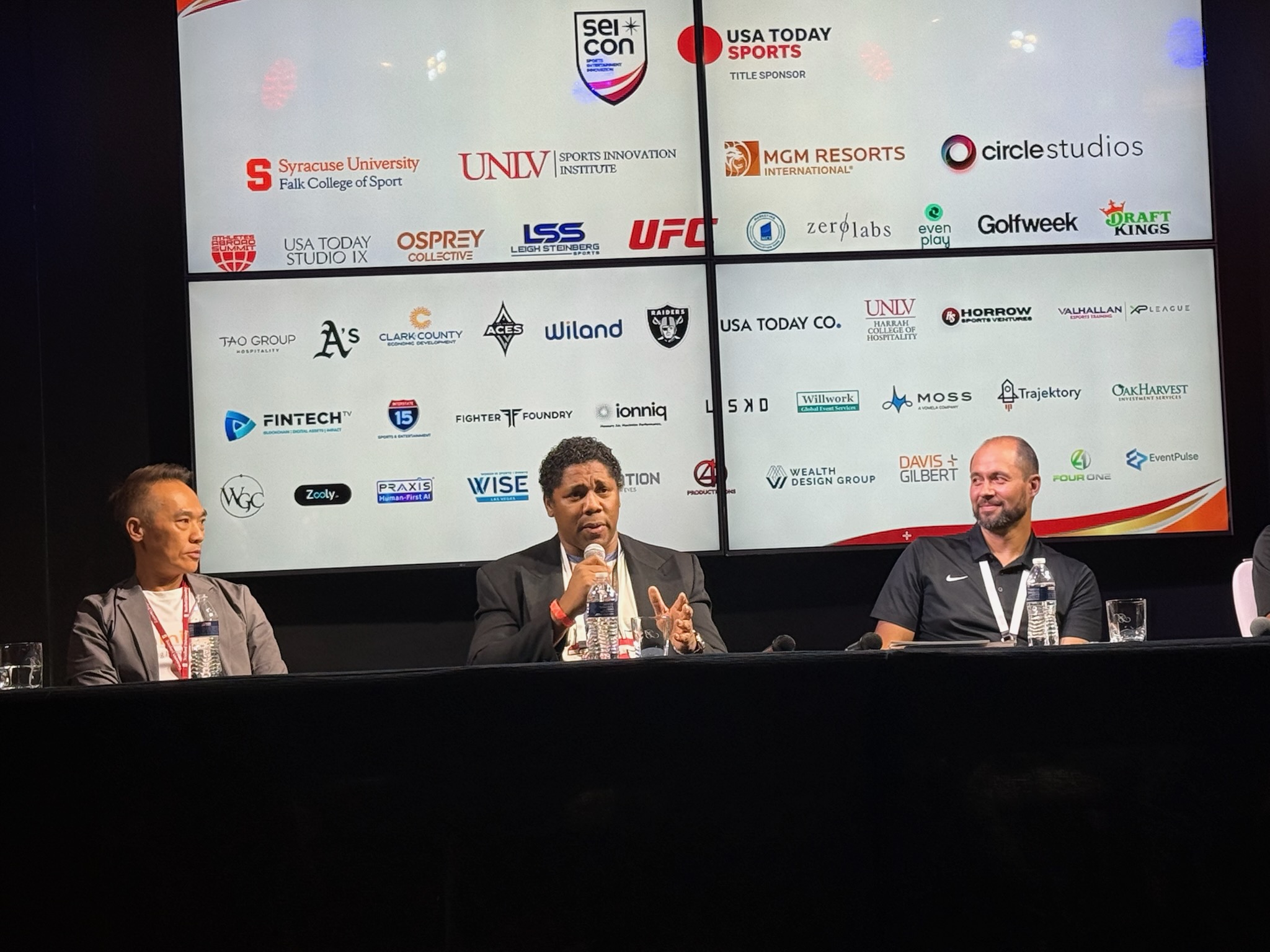 Three people seated at a panel discussion with a large screen behind them displaying various logos and sponsors.