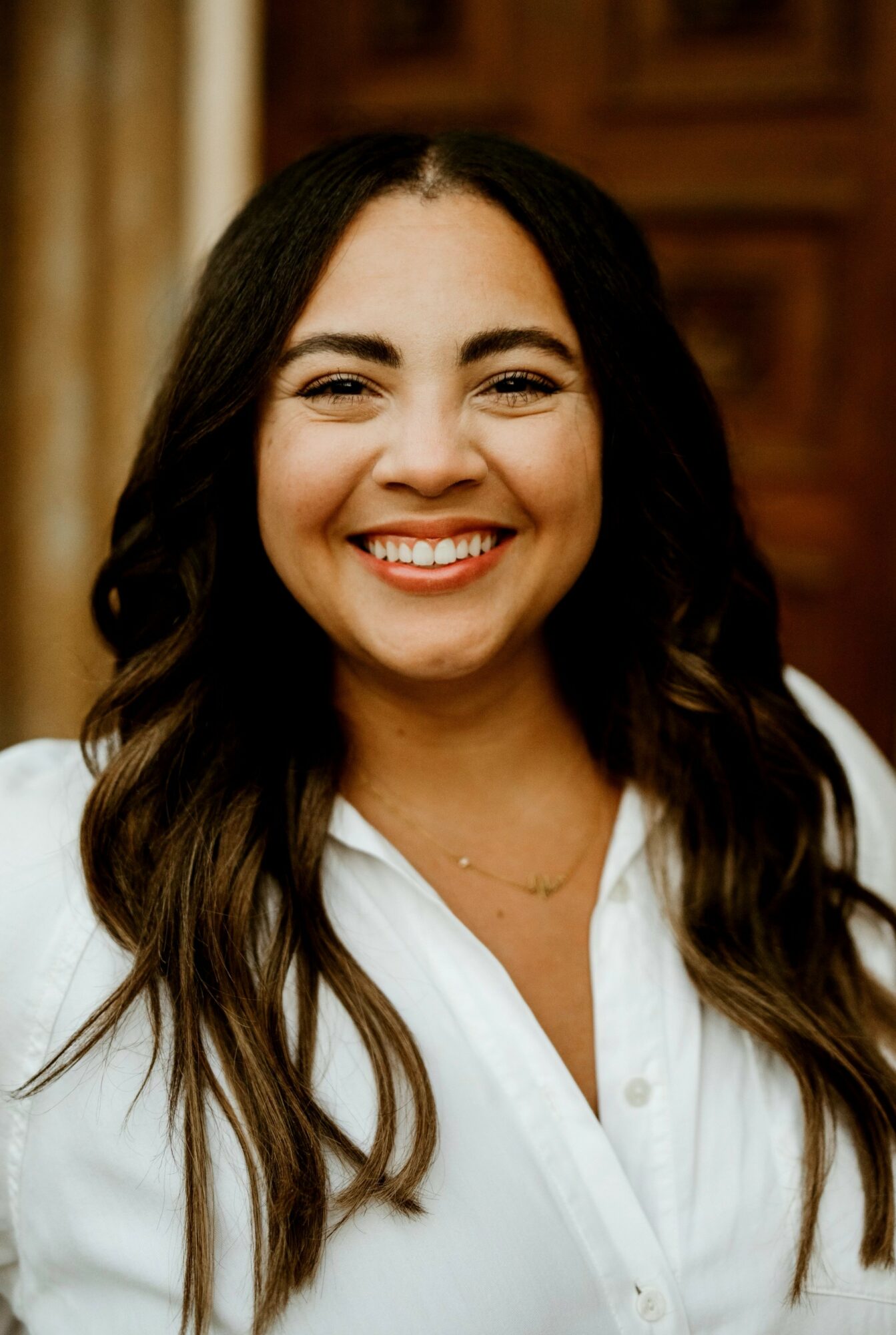 Smiling woman with long dark hair wearing a white shirt, indoors with wooden background.
