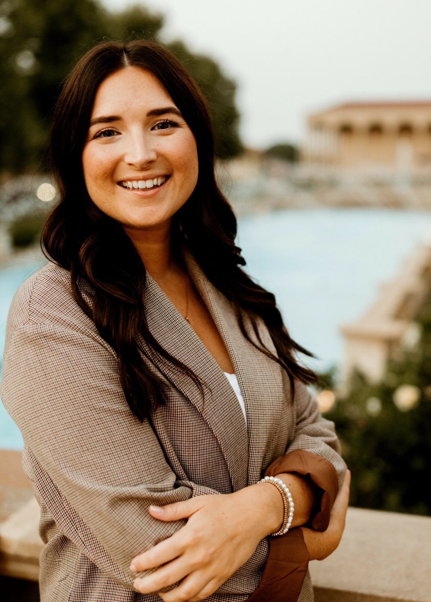 Smiling woman with long dark hair, wearing a beige blazer, standing outdoors near water and trees.