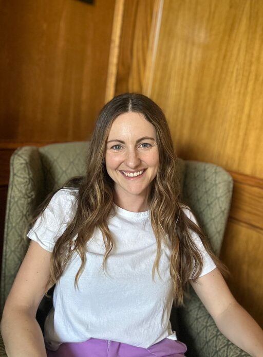 Smiling woman with long wavy hair sitting in a green armchair against wooden paneling background.