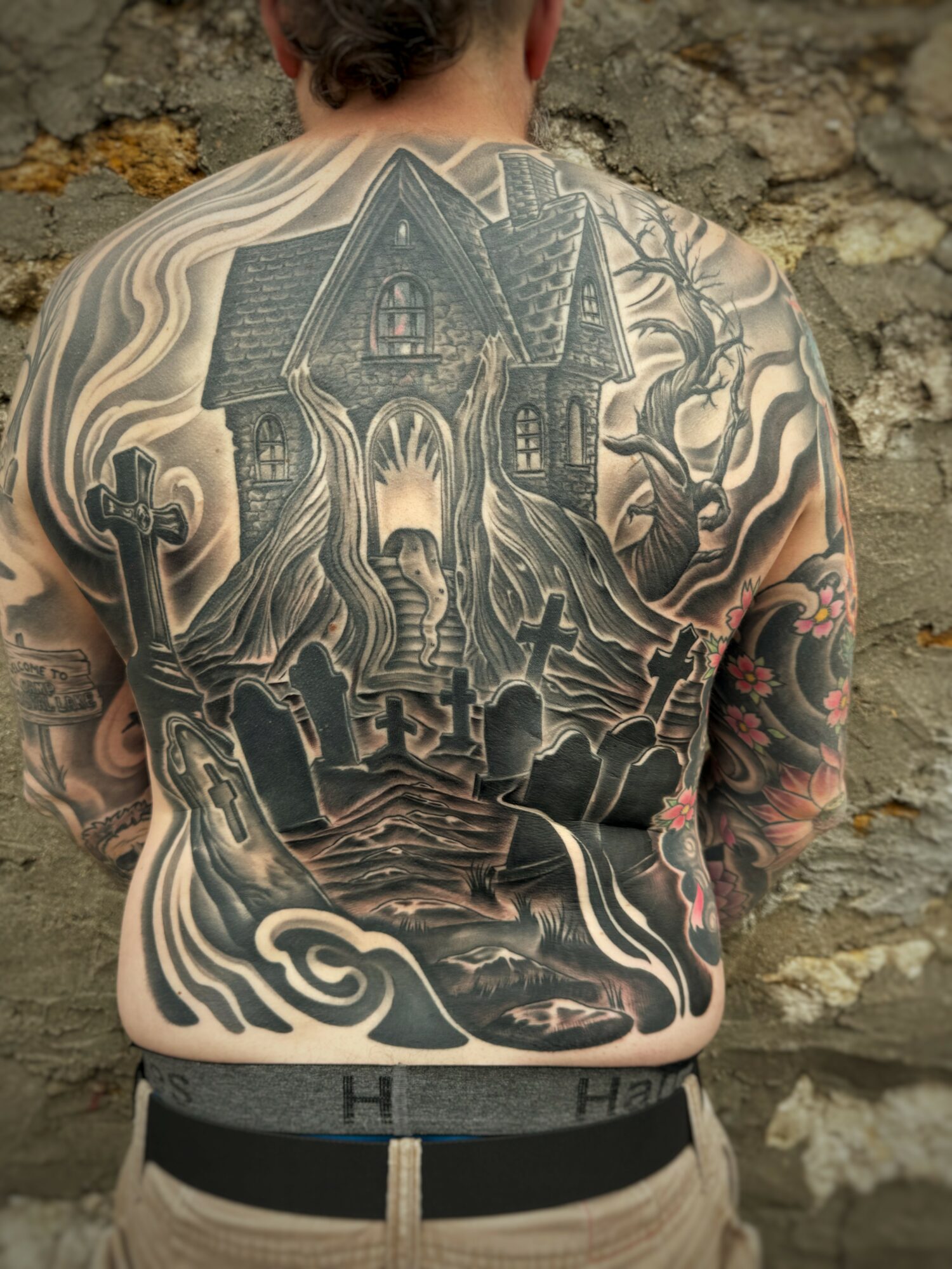 Back tattoo of a haunted house with trees, crosses, and swirling patterns, covering the person's back and shoulders.