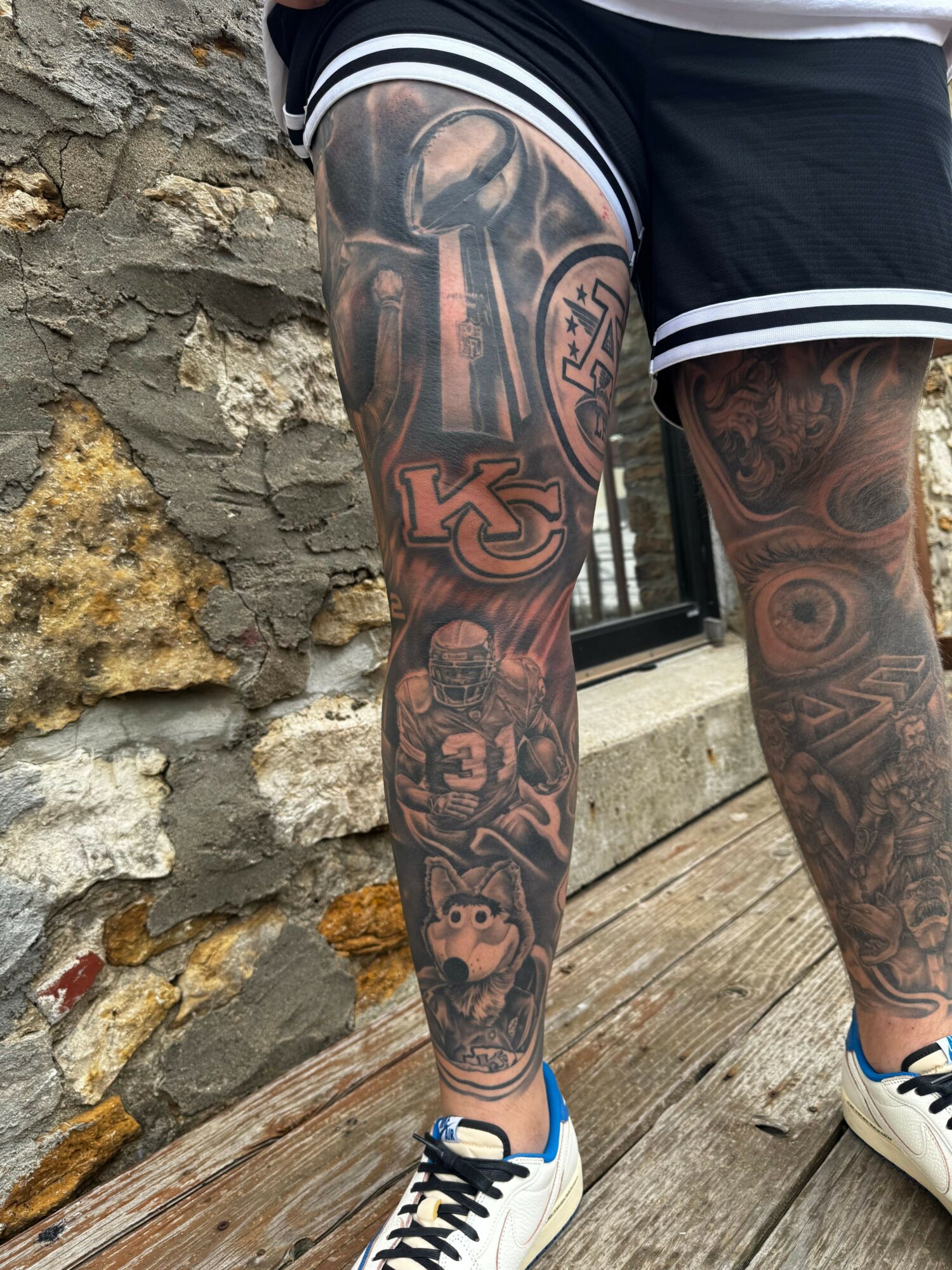 Tattooed legs with shorts and sneakers resting on a wooden surface, with a rocky ground below.