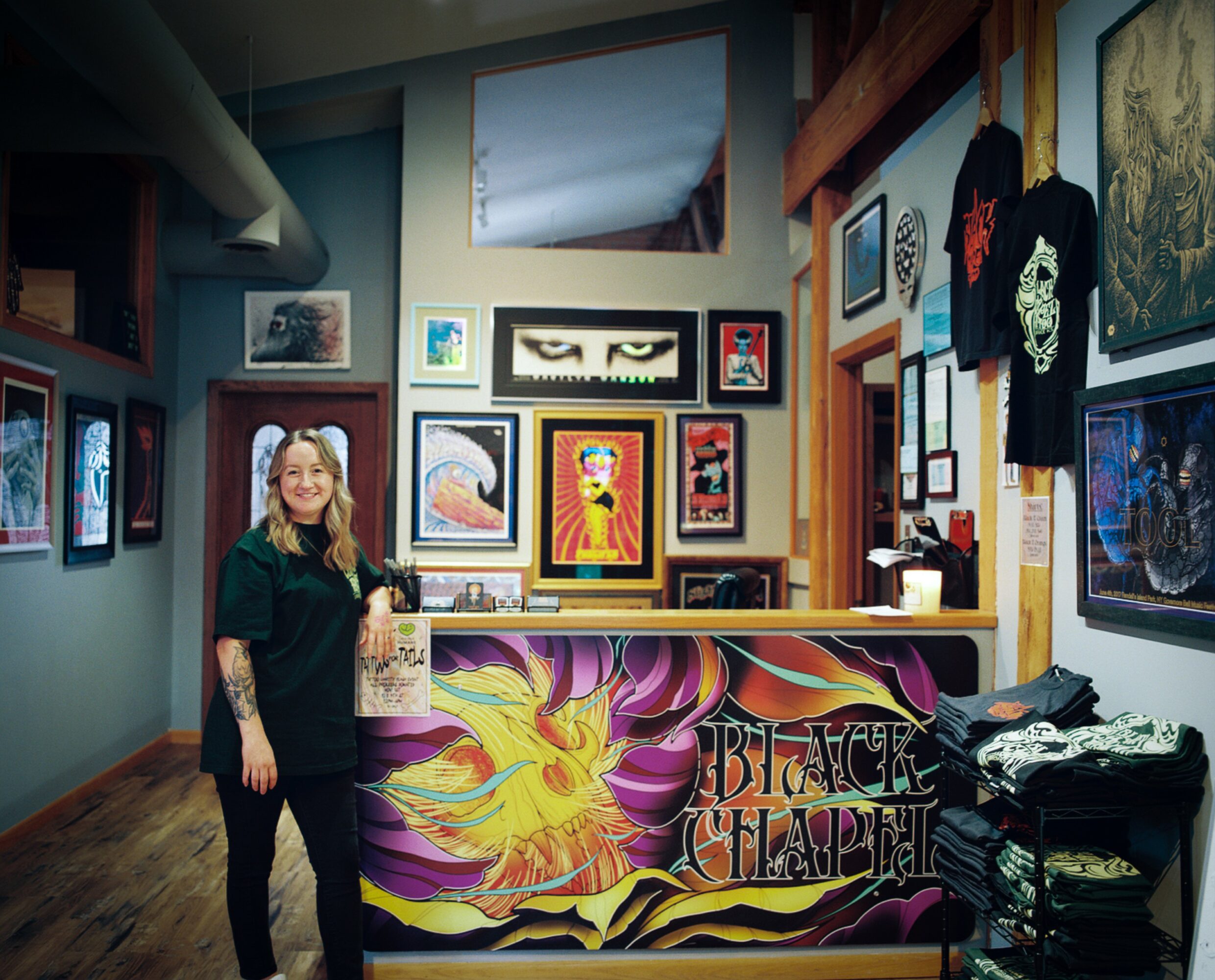 Woman standing in art gallery with colorful paintings and a decorated counter, wooden beams, and framed artwork on walls.