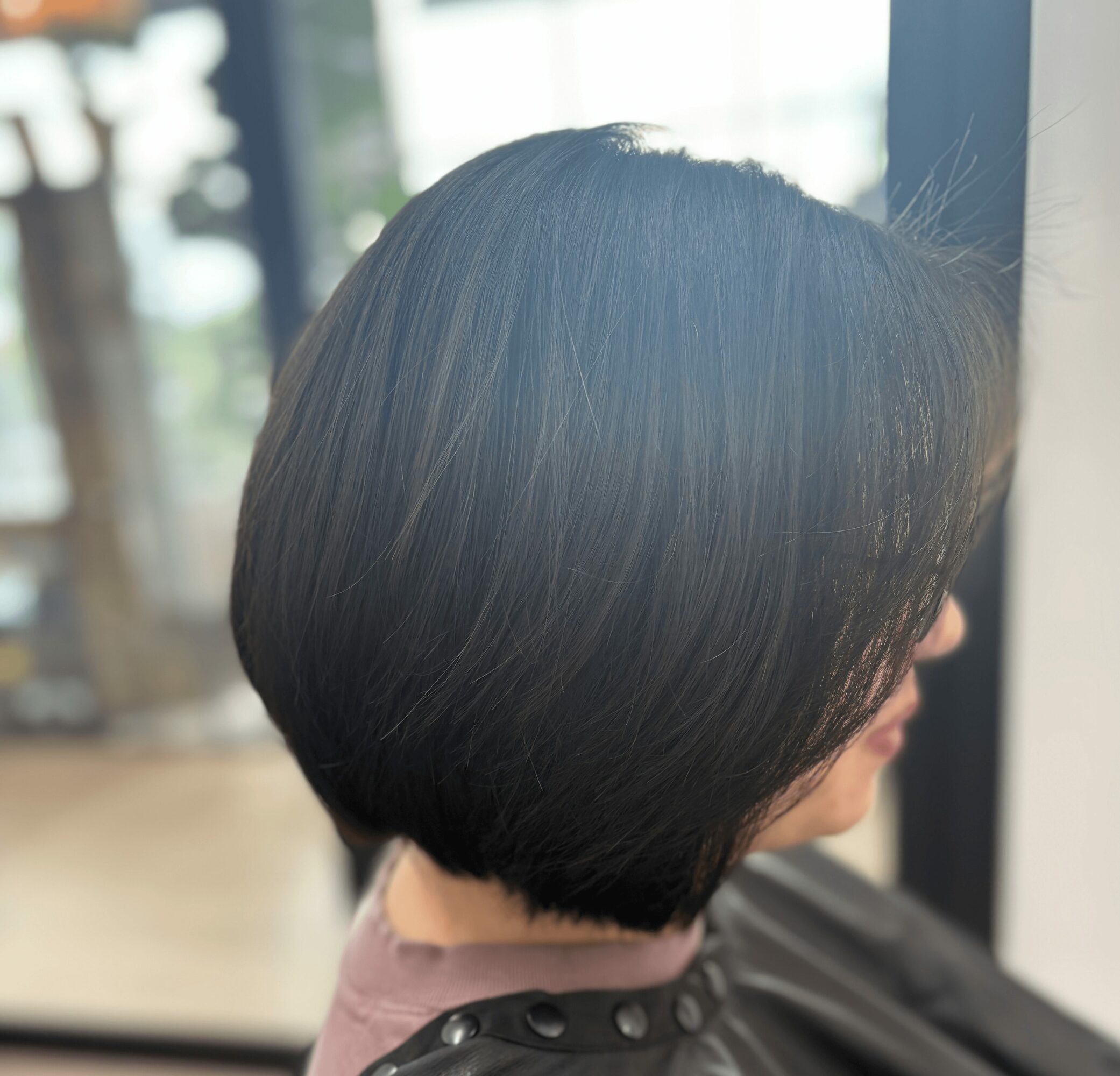 Side view of a person with a short, dark, straight bob haircut, sitting near a window.