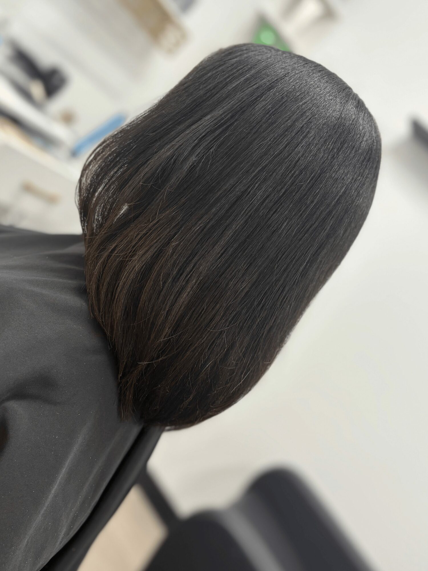 Person with straight, dark hair sitting in a salon chair, facing away, with a blurred background.