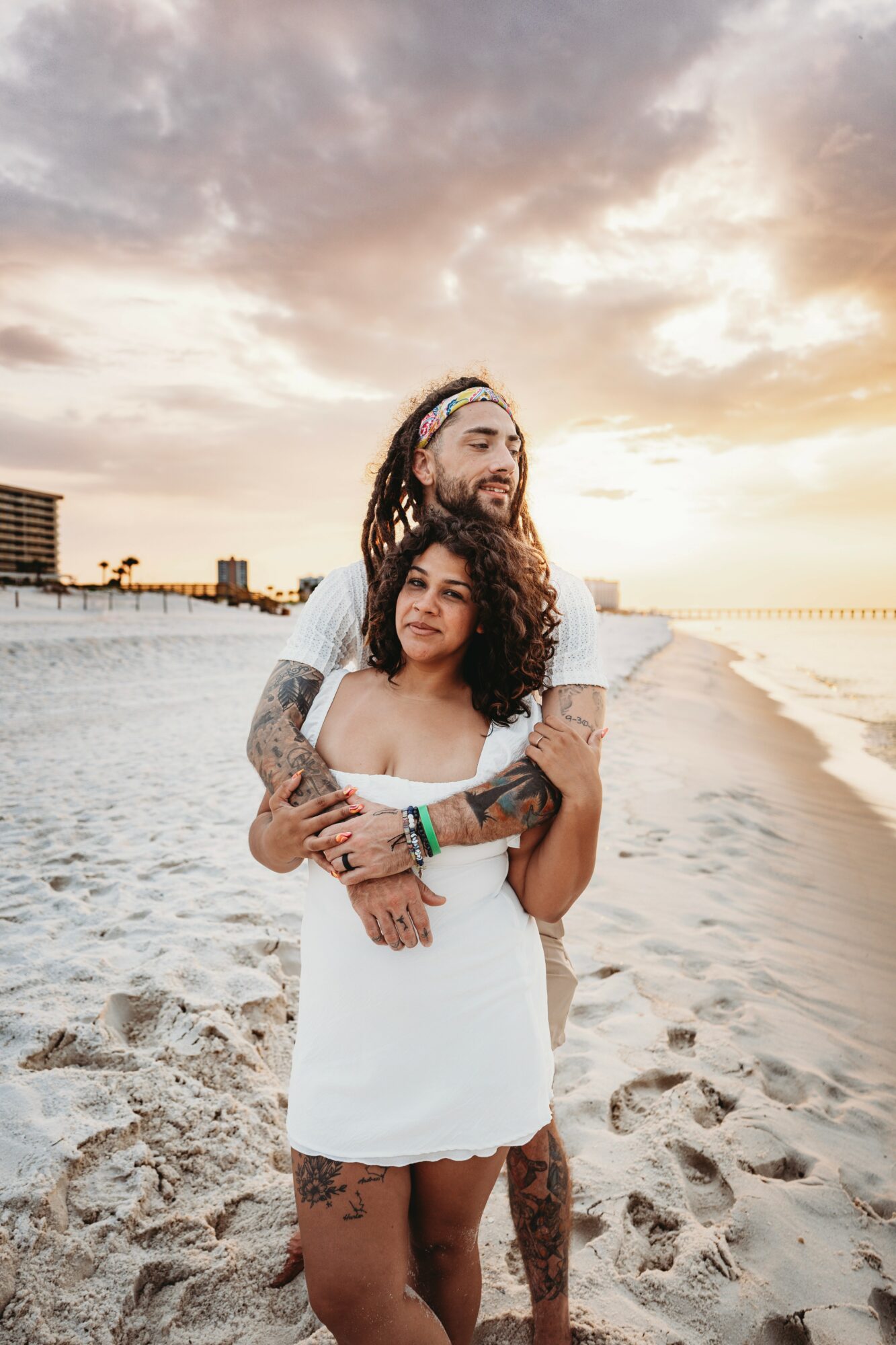 A couple stands on a beach during sunset, embracing each other with buildings in the background.