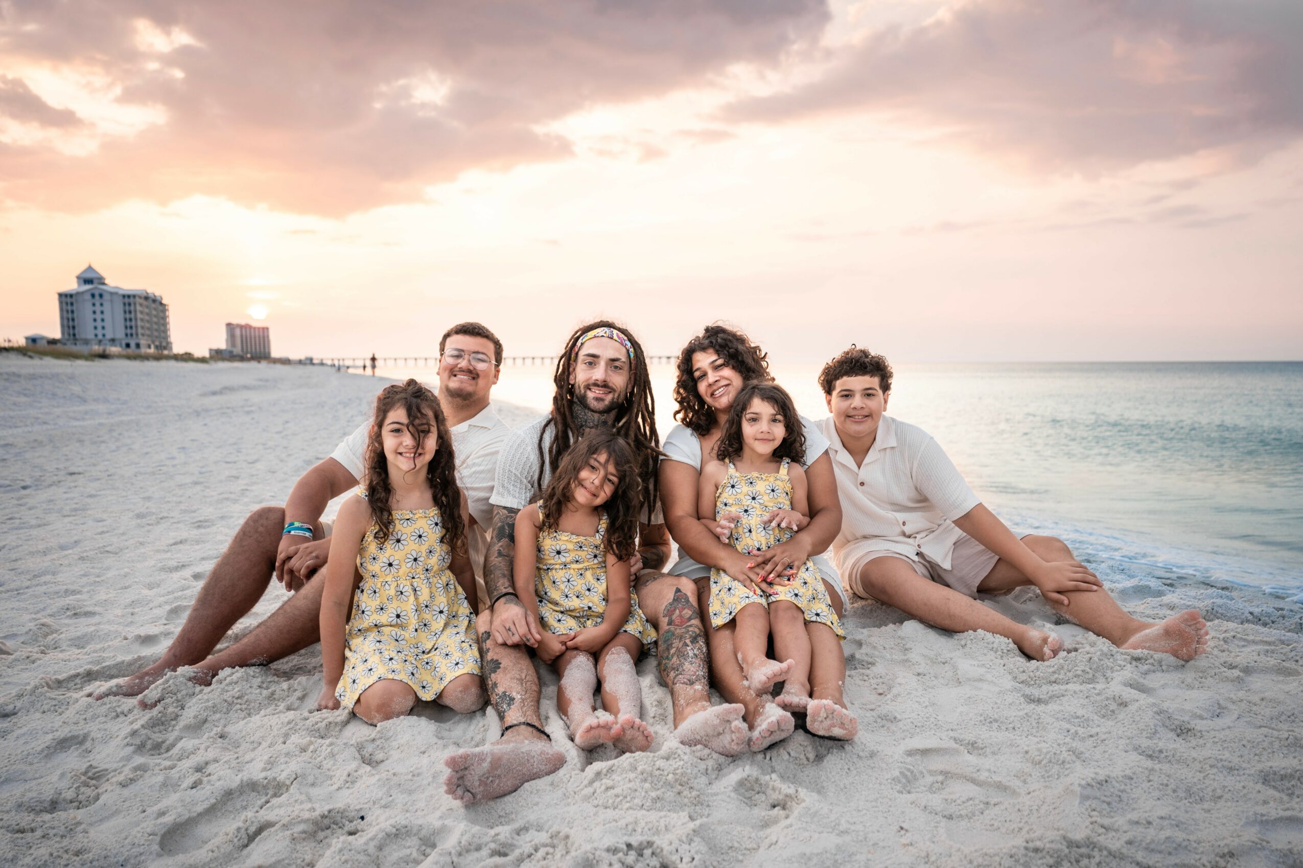 Group of seven people sitting on the beach at sunset, with buildings in the background, smiling and relaxing.
