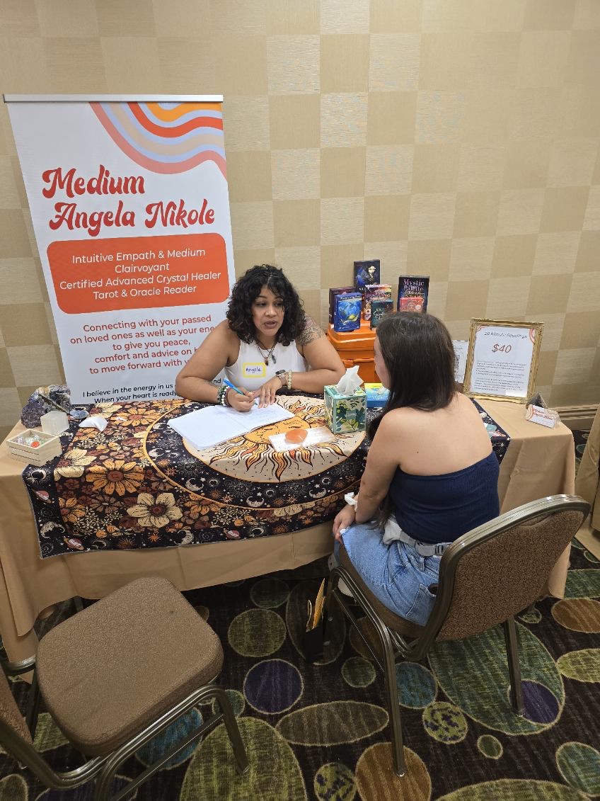 Two women sit at a table with promotional materials, a sign, and books, engaging in conversation at an event.
