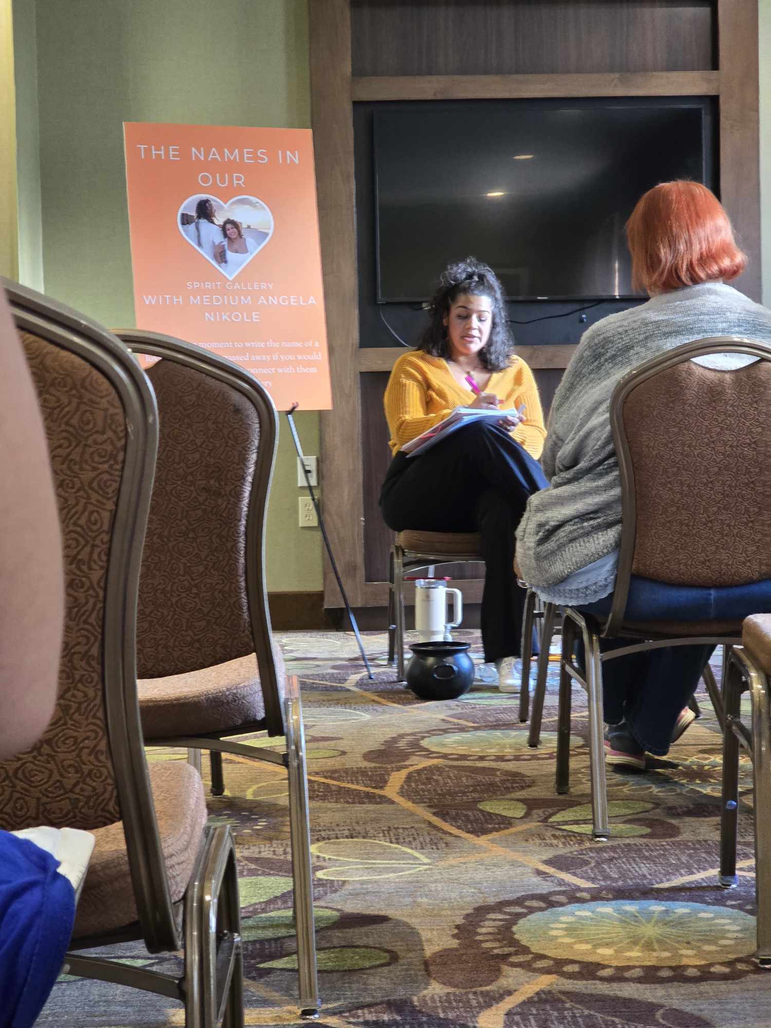 Two women seated and talking in a room with chairs, a large TV, and a poster on the wall.